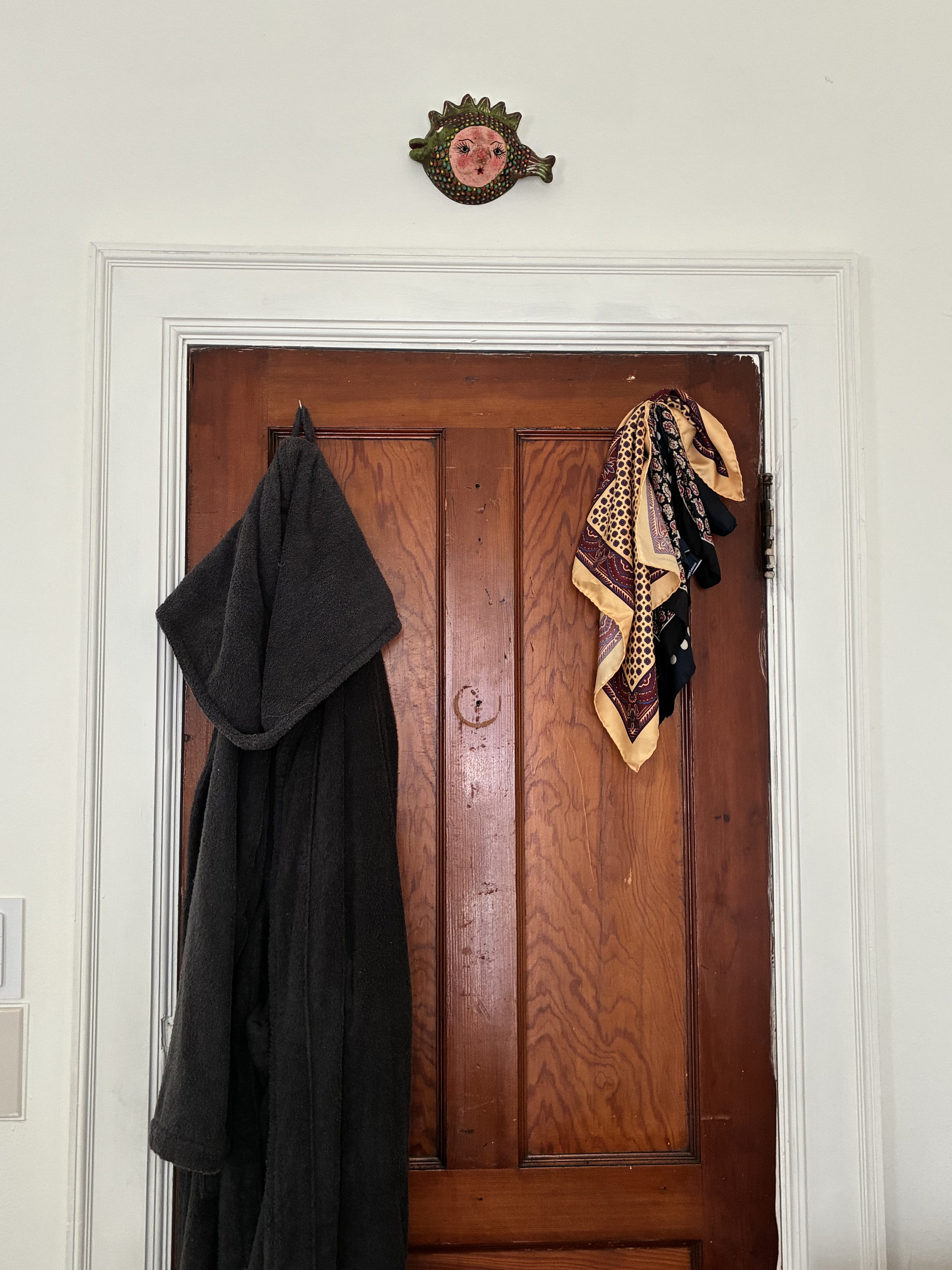 A wooden door with a dark brown color and visible wood grain. There are two hooks on the door, one with a black towel hanging on it and the other holding a colorful scarf. Above the door, there is a decorative metal wall art piece resembling a fish with a face, painted in bright colors and with floral and fantasy design elements.