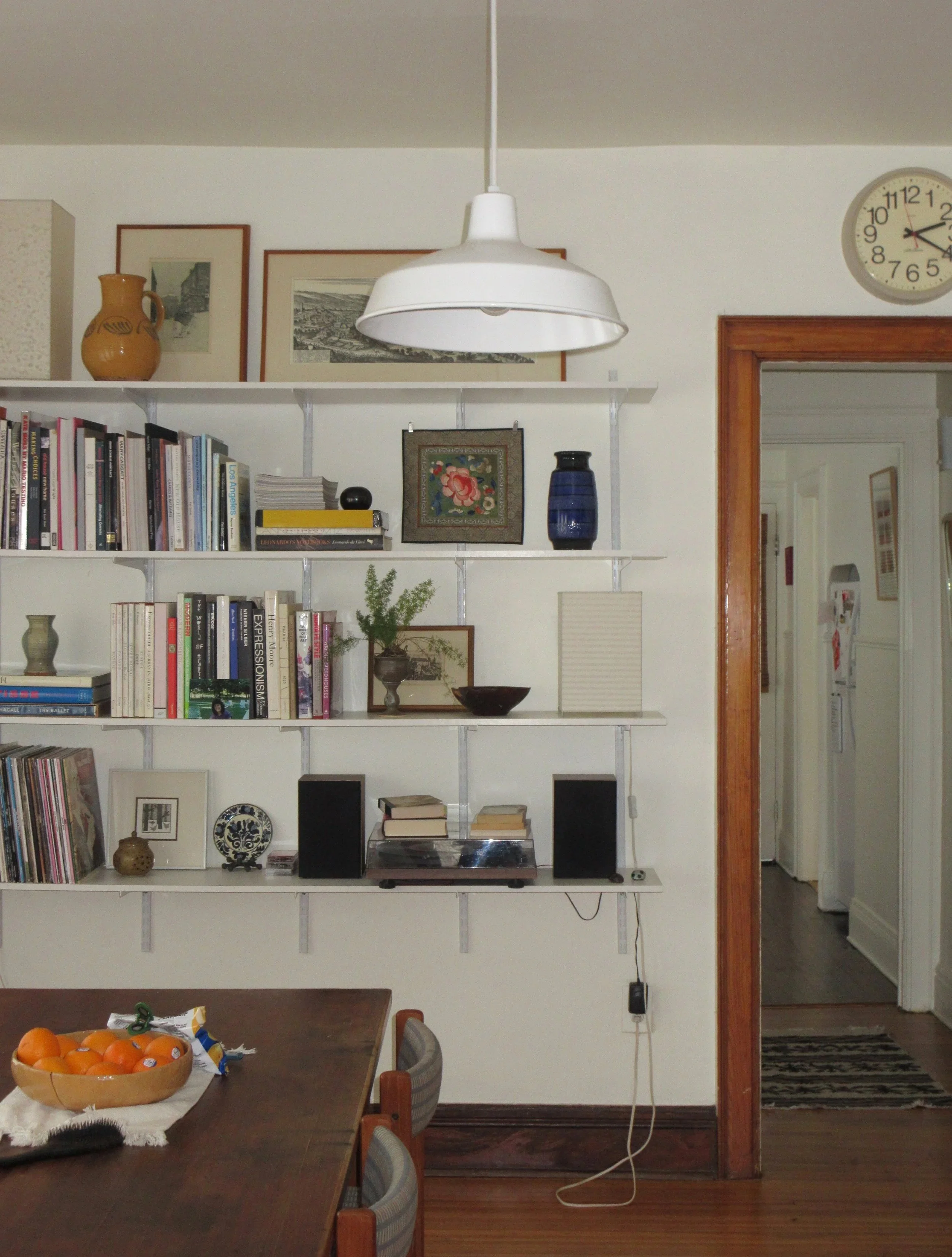 A room with a wall-mounted white bookshelf filled with books, decorative objects, and framed pictures. A wooden dining table with a fruit bowl and some snacks is in the foreground. A white pendant lamp hangs from the ceiling, and a clock shows the time as 12:16.