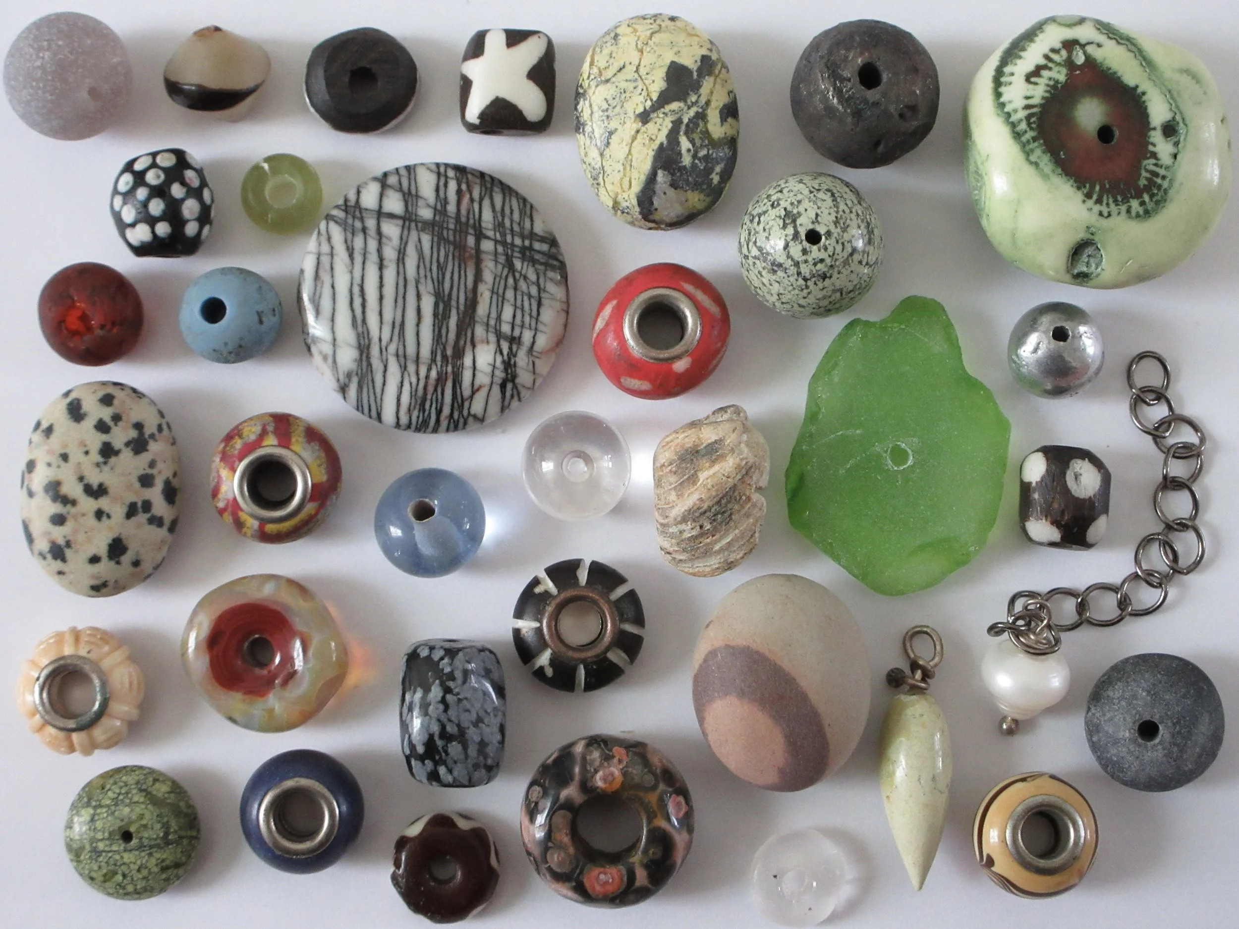 A collection of various beads made from different materials, colors, and patterns, including glass, stone, and ceramic, some with holes for stringing, arranged on a white background.