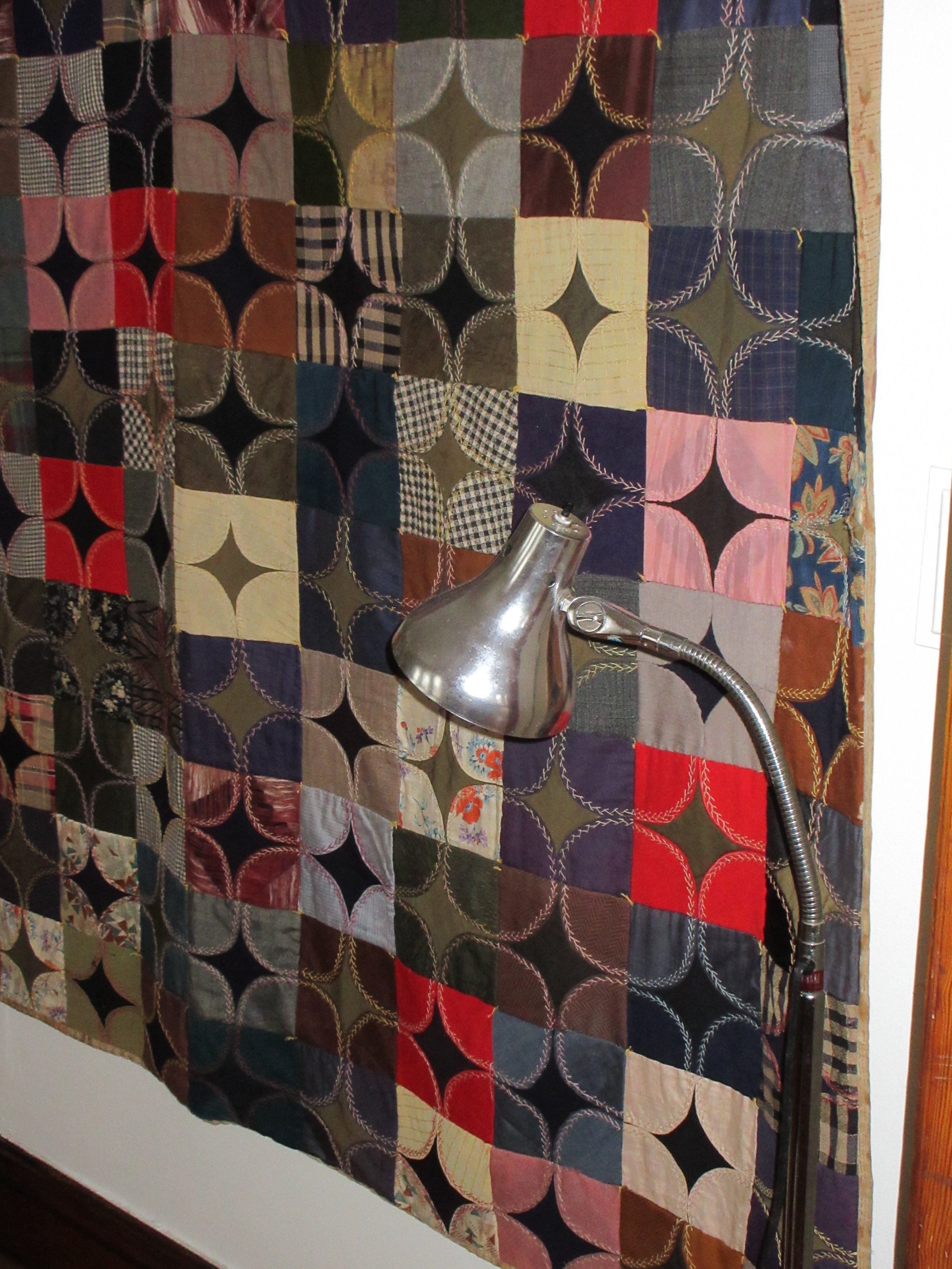 Colorful quilt with various geometric and patterned squares hanging on a wall, with a silver adjustable desk lamp attached to a pole in front of it.