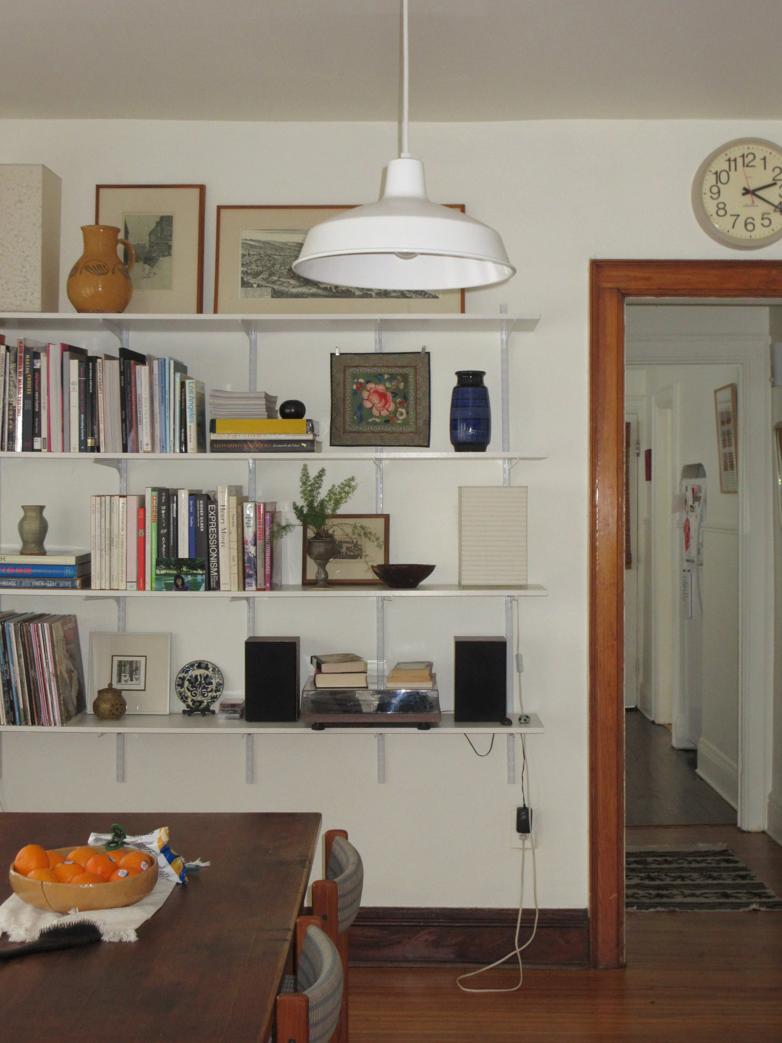 Interior of a dining room with a wooden table, a shelf filled with books, decorations, and a turntable. A white pendant lamp hangs from the ceiling, and a wall clock is visible in the corner.