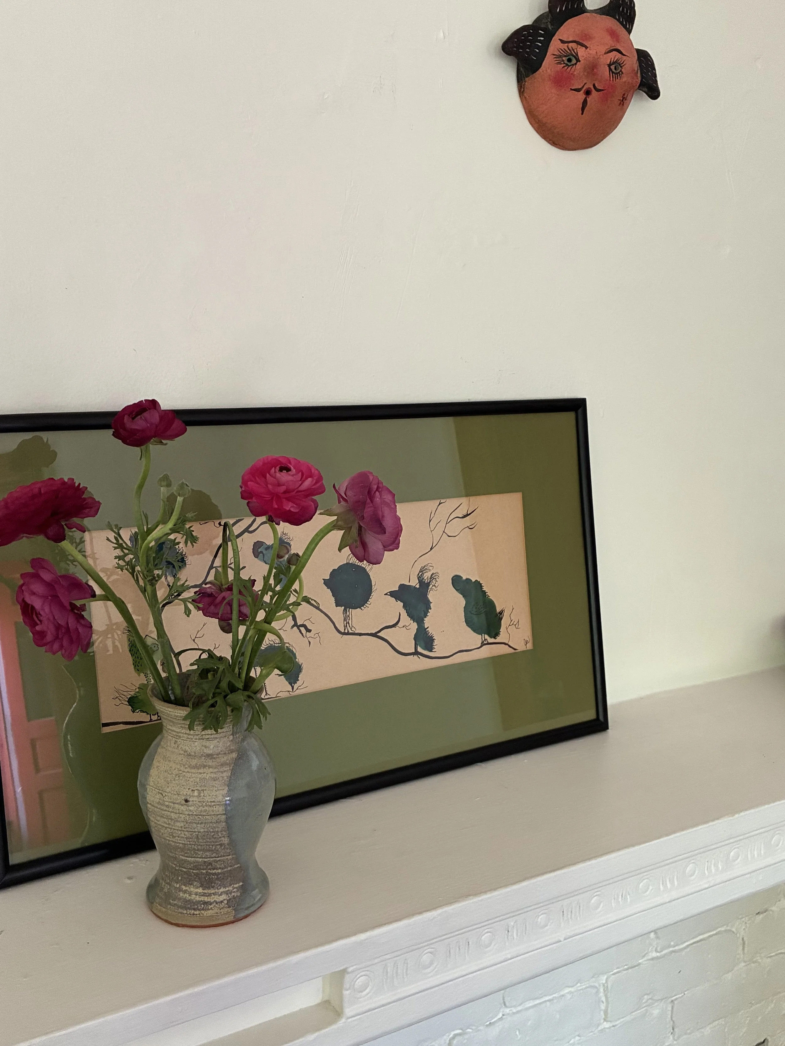 A vase with pink flowers sitting on a white mantel, with a framed art piece and a wall decoration depicting a sun with a face hanging above.