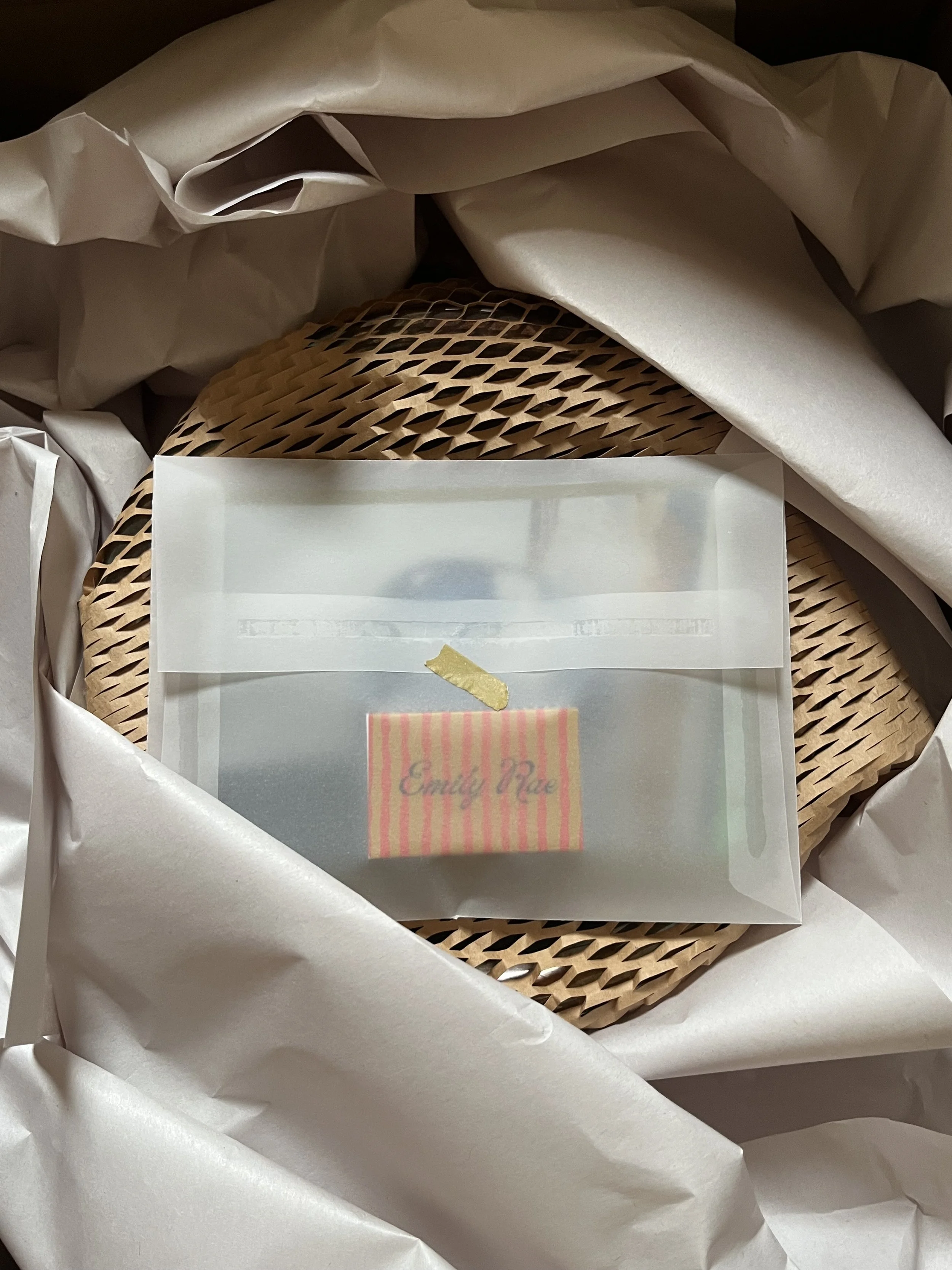 Wrapped gift with a round honeycomb-patterned box inside white tissue paper, with a small pink and yellow striped card labeled 'Emily Mae' inside a transparent plastic pouch.
