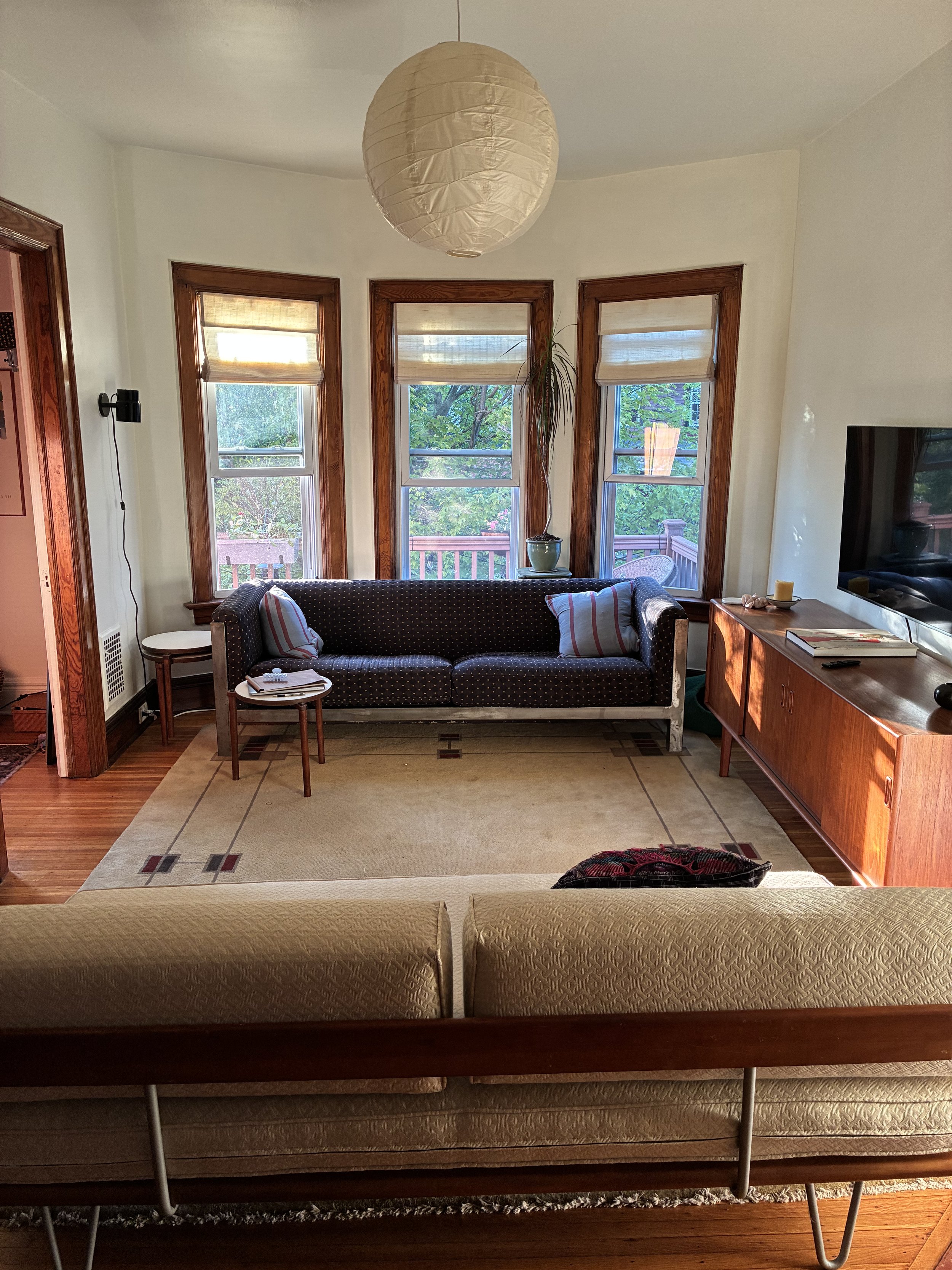 Living room with three large front windows, a dark patterned sofa with throw pillows, a small side table, a wooden sideboard with candles, a large wall-mounted TV, and beige area rug, with sunlight illuminating the room.