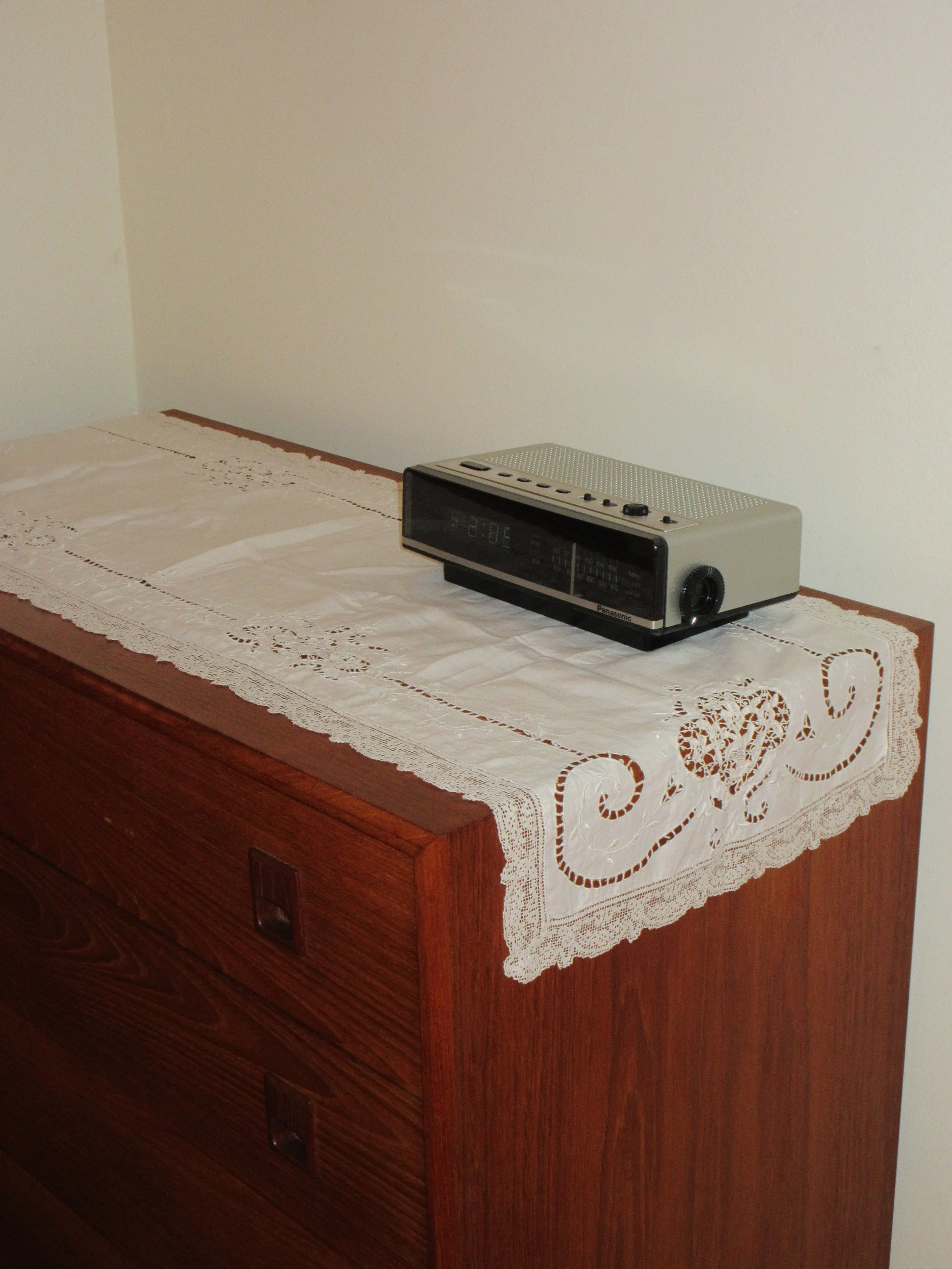 A vintage alarm clock sitting on a wooden dresser covered with a lace doily.