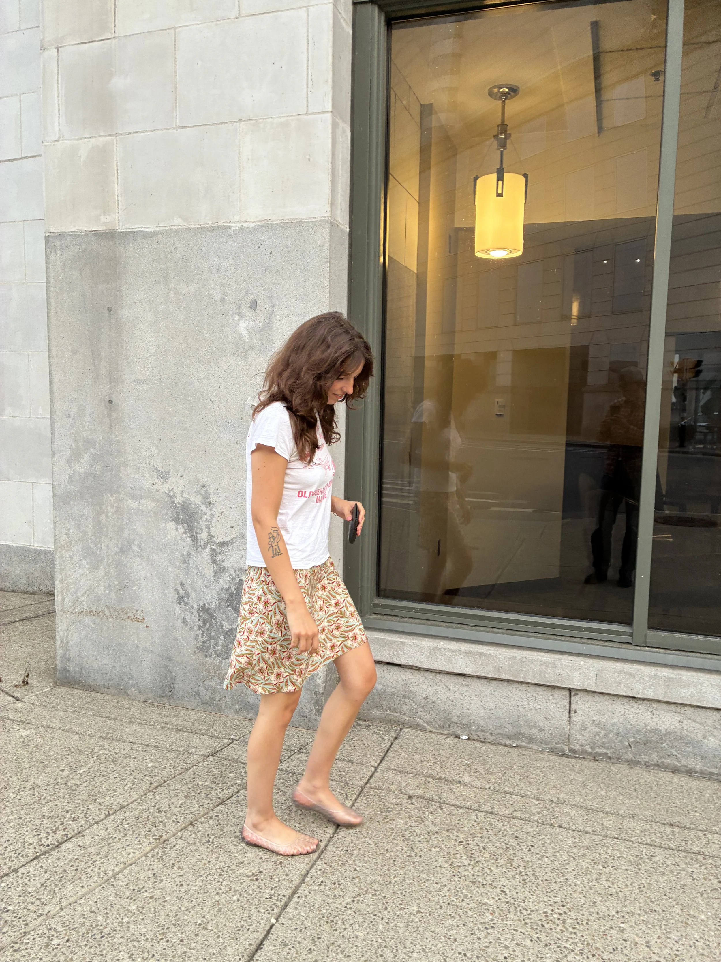 A woman with brown hair, wearing a white t-shirt, a patterned skirt, and clear shoes, is standing outdoors on a sidewalk in front of a building with a large glass window and concrete walls.