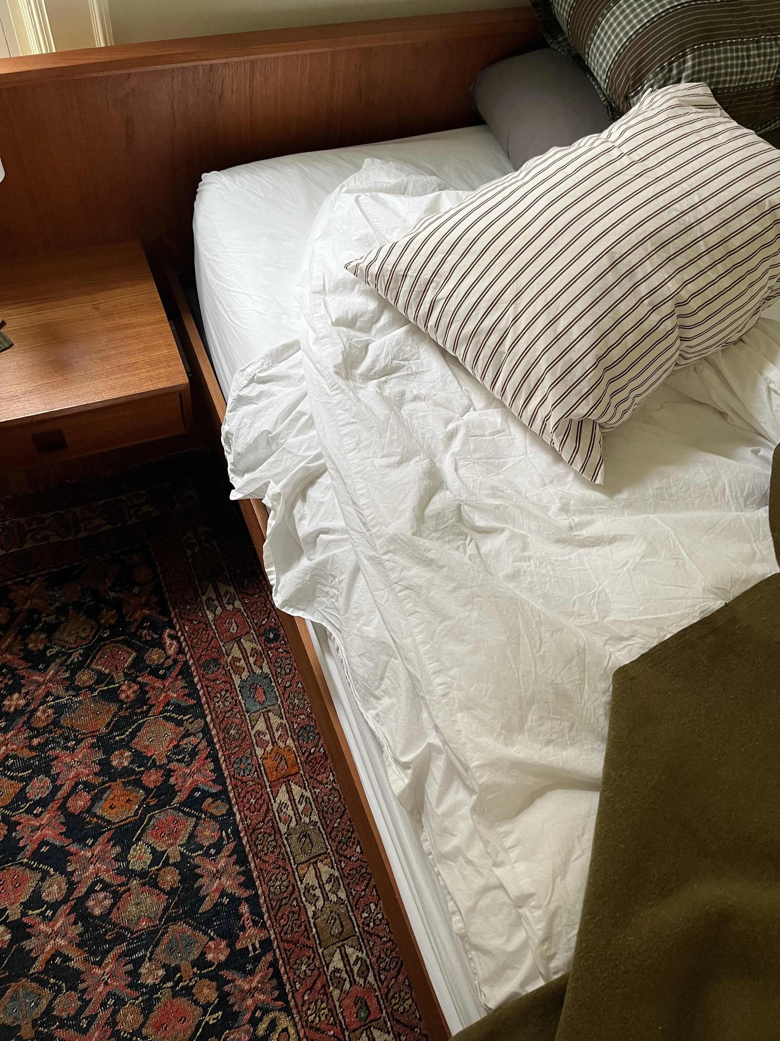 A neatly made bed with white linens, a striped pillow, a plain pillow, and a dark checkered pillow, with a wooden headboard and a nightstand beside it. The bed is next to a richly patterned, multicolored rug.