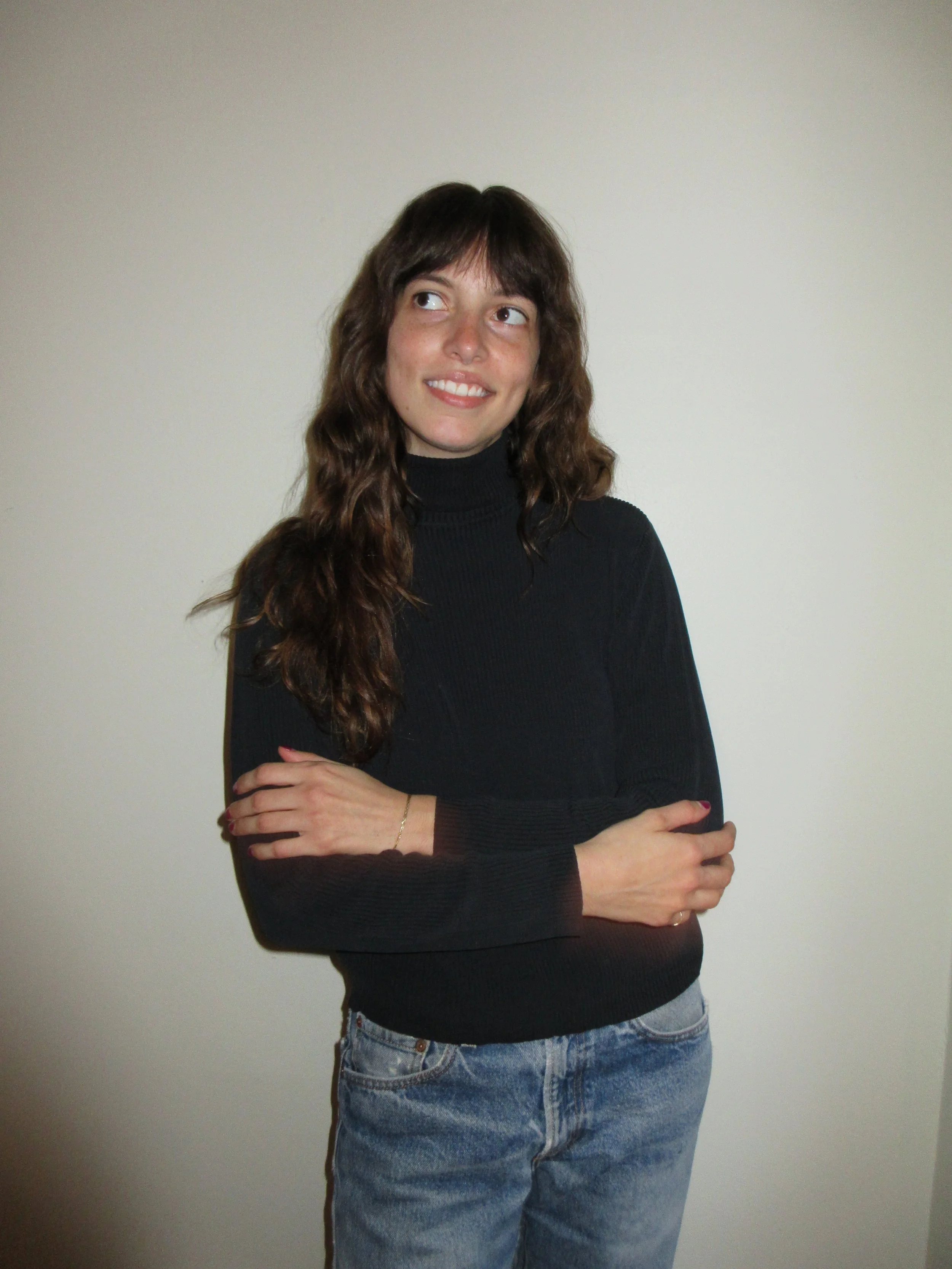 A woman with long wavy brown hair wearing a black turtleneck sweater and light blue jeans, standing against a plain wall. She is crossing her arms and looking to her left with a slight smile.