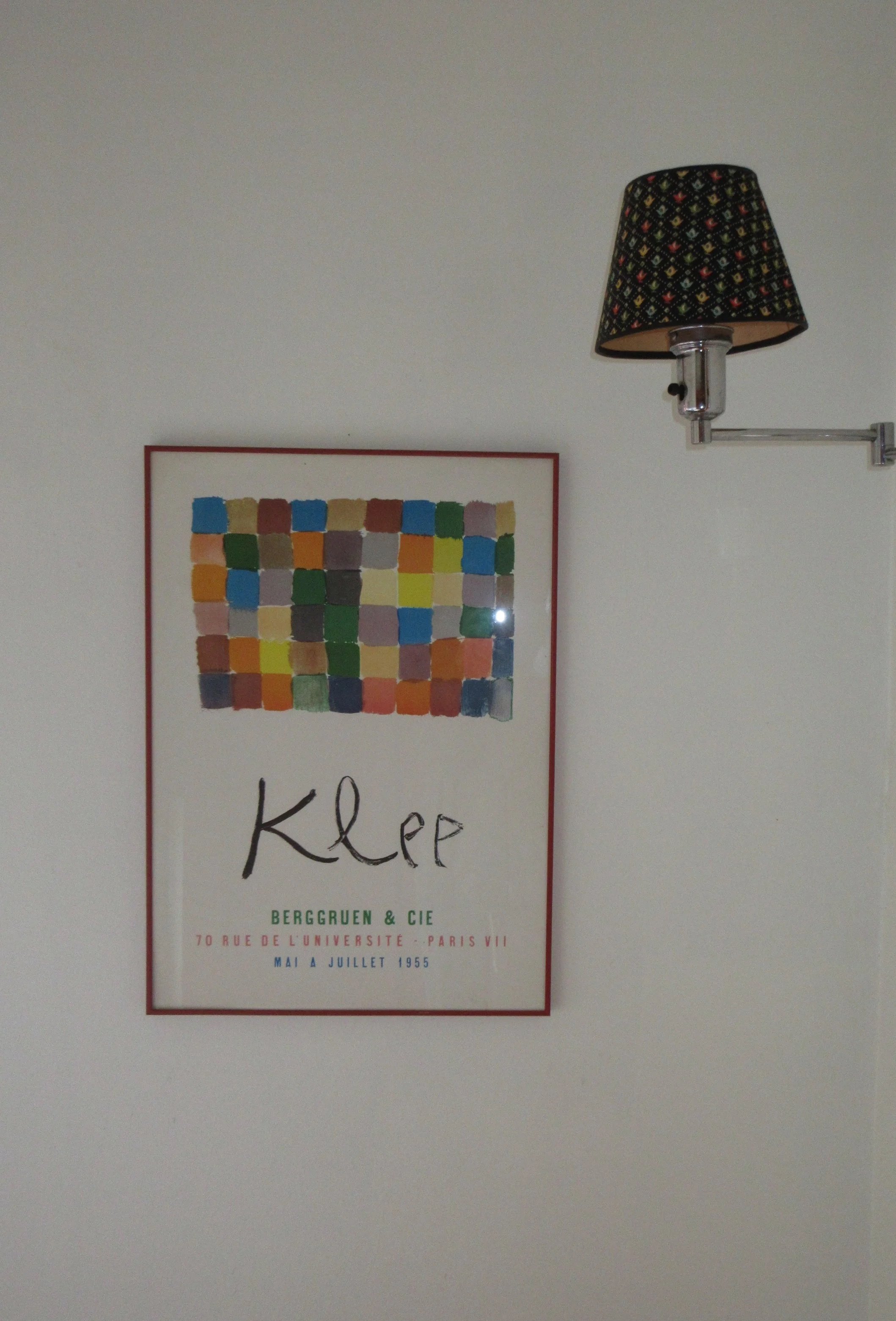 A framed poster on a white wall featuring a grid of colorful squares and the word 'Klep' written beneath. To the right of the poster is a wall-mounted lamp with a black lampshade decorated with small multicolored dots.