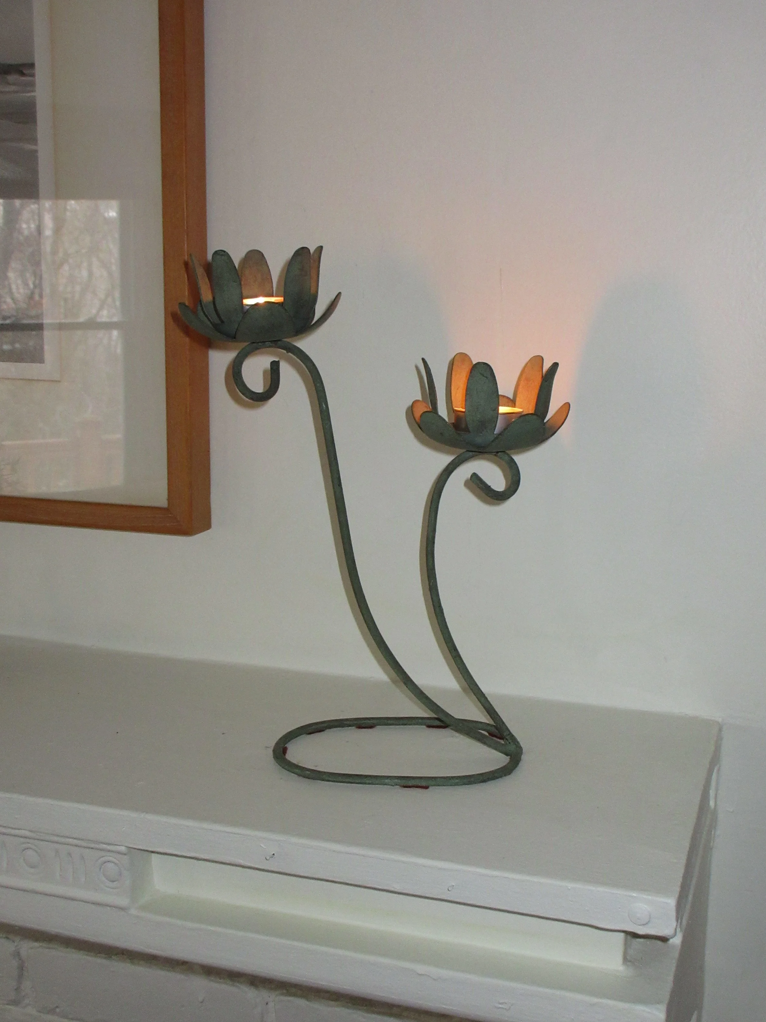 A metal candle holder with two flower-shaped holders, lit candles inside, positioned on a white surface against a light-colored wall.