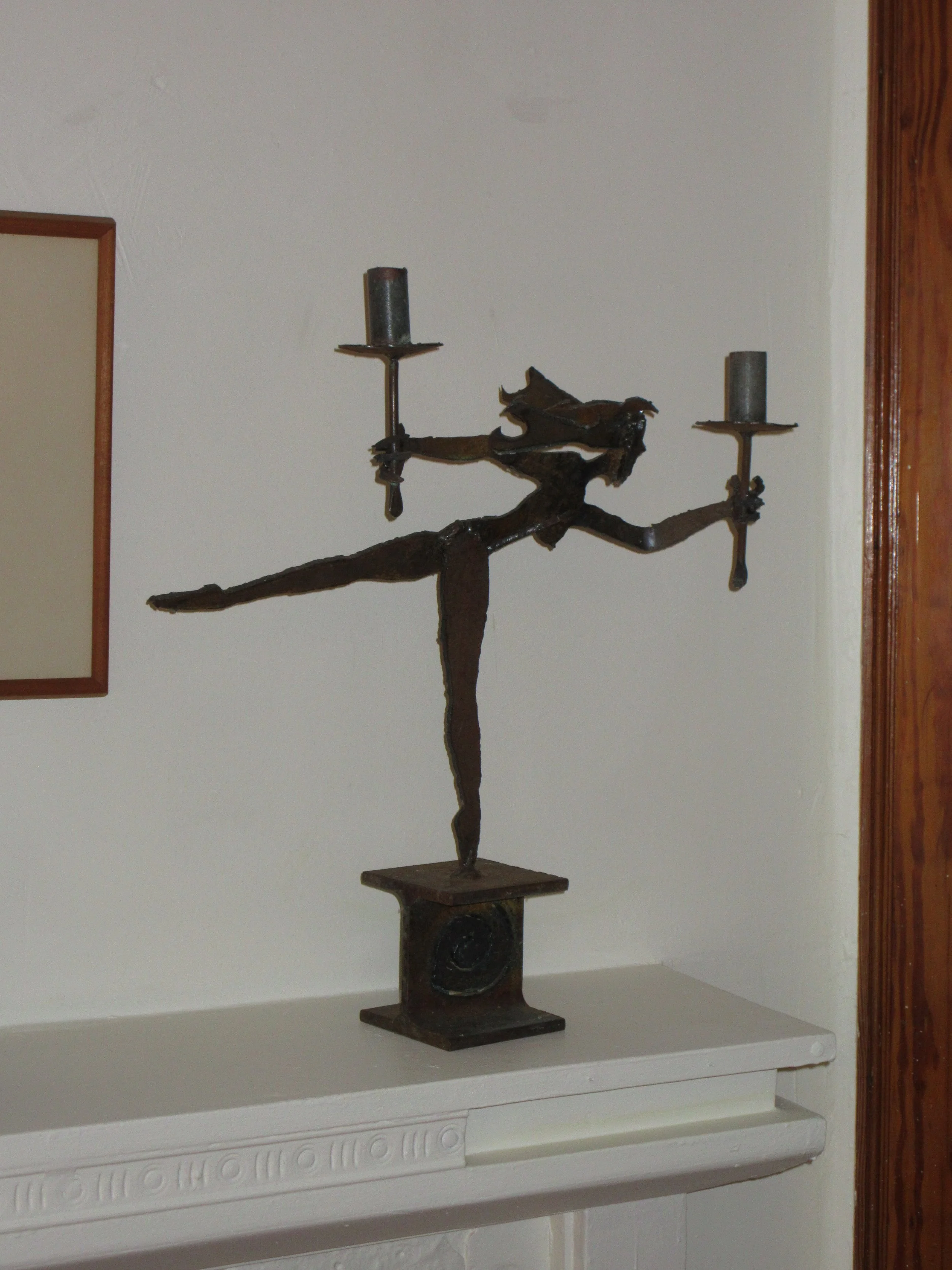 Metal sculpture of a woman dancing ballet, holding candles in each hand, placed on a white mantelpiece against a beige wall.