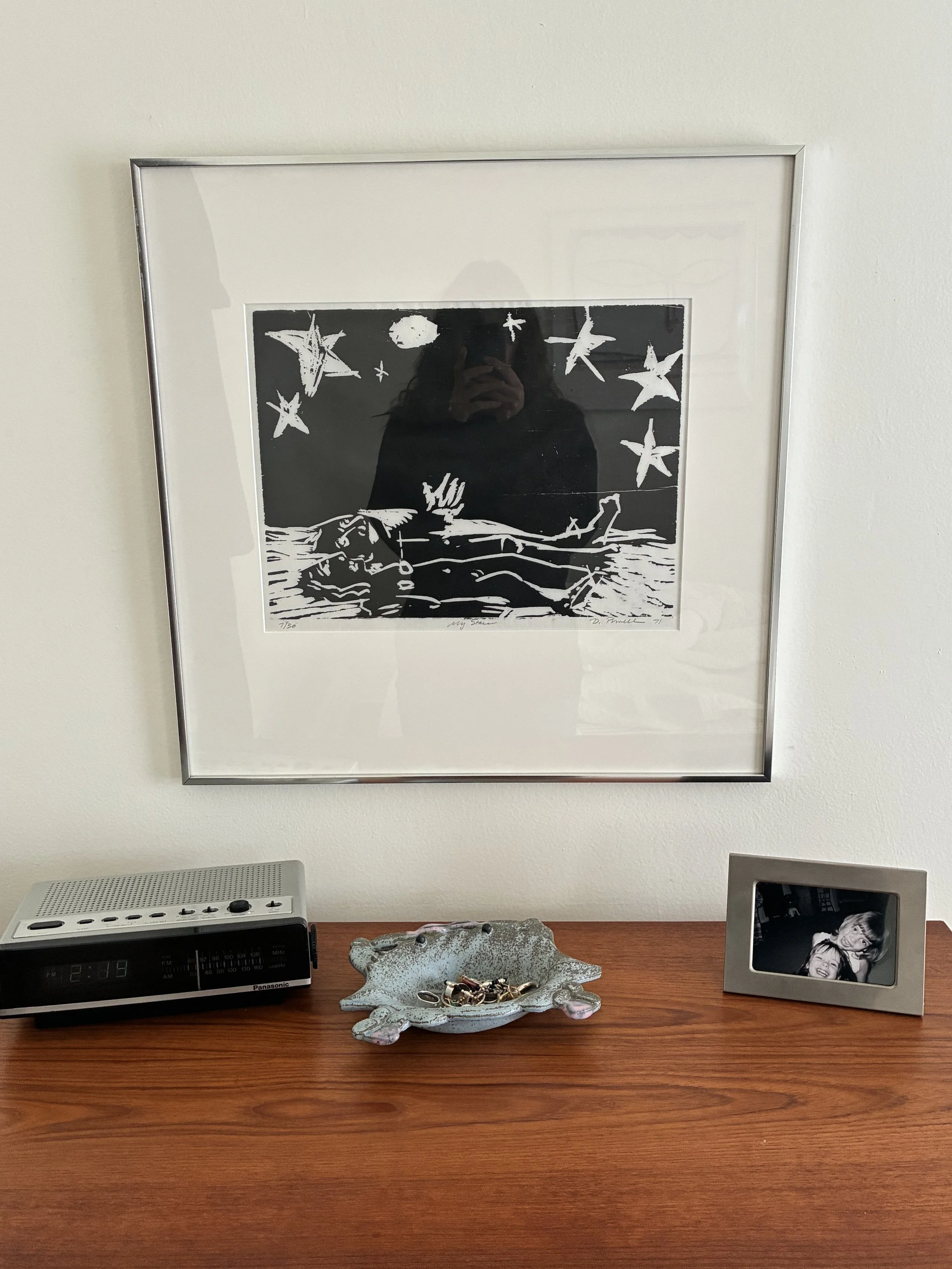 A framed black and white art print hanging on a wall above a wooden table. The table holds a digital alarm clock, a decorative dish with jewelry, and a picture frame with a photo of two women.