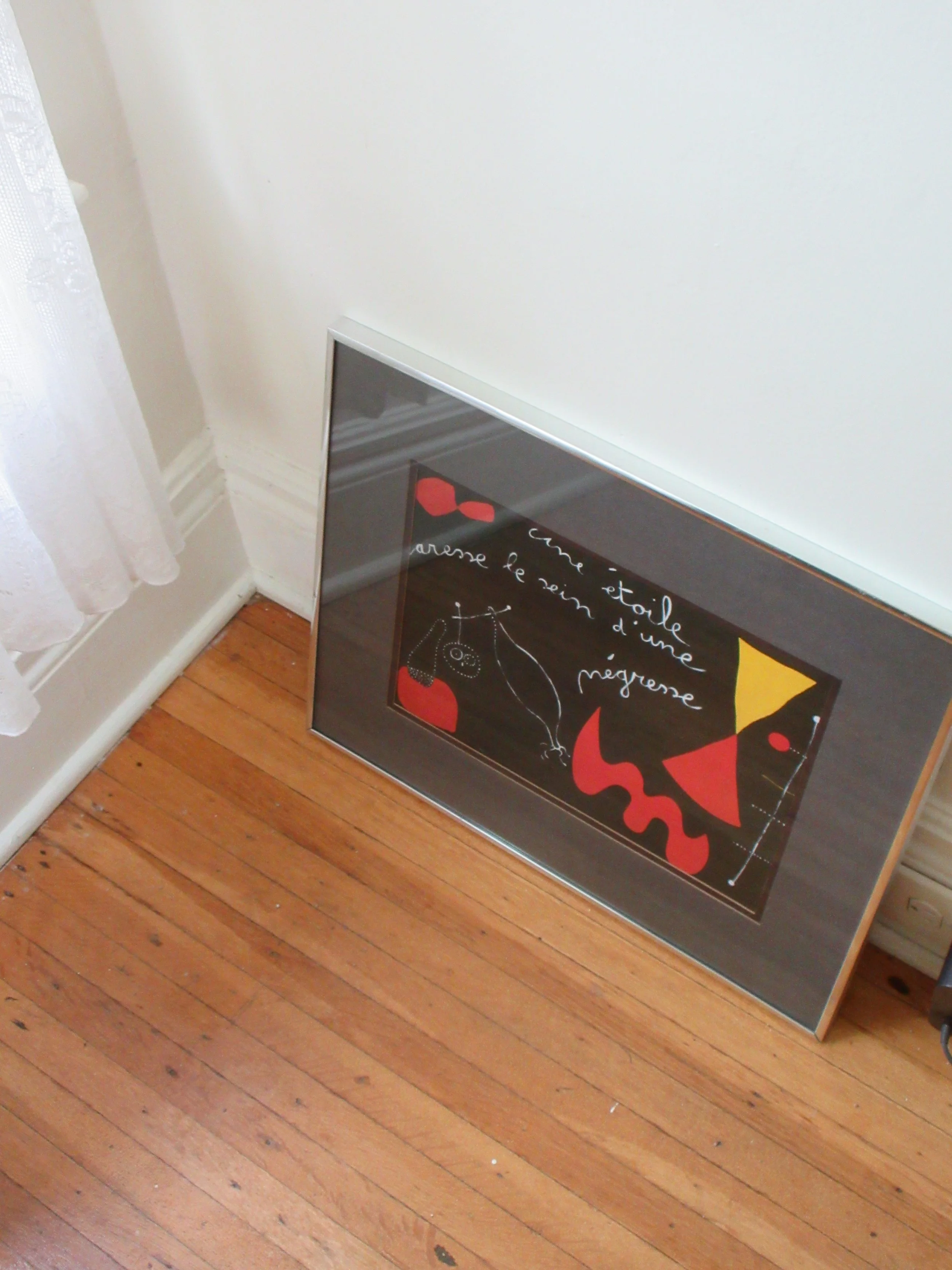 Framed art piece leaning against white wall near window with lace curtain, on wooden floor.