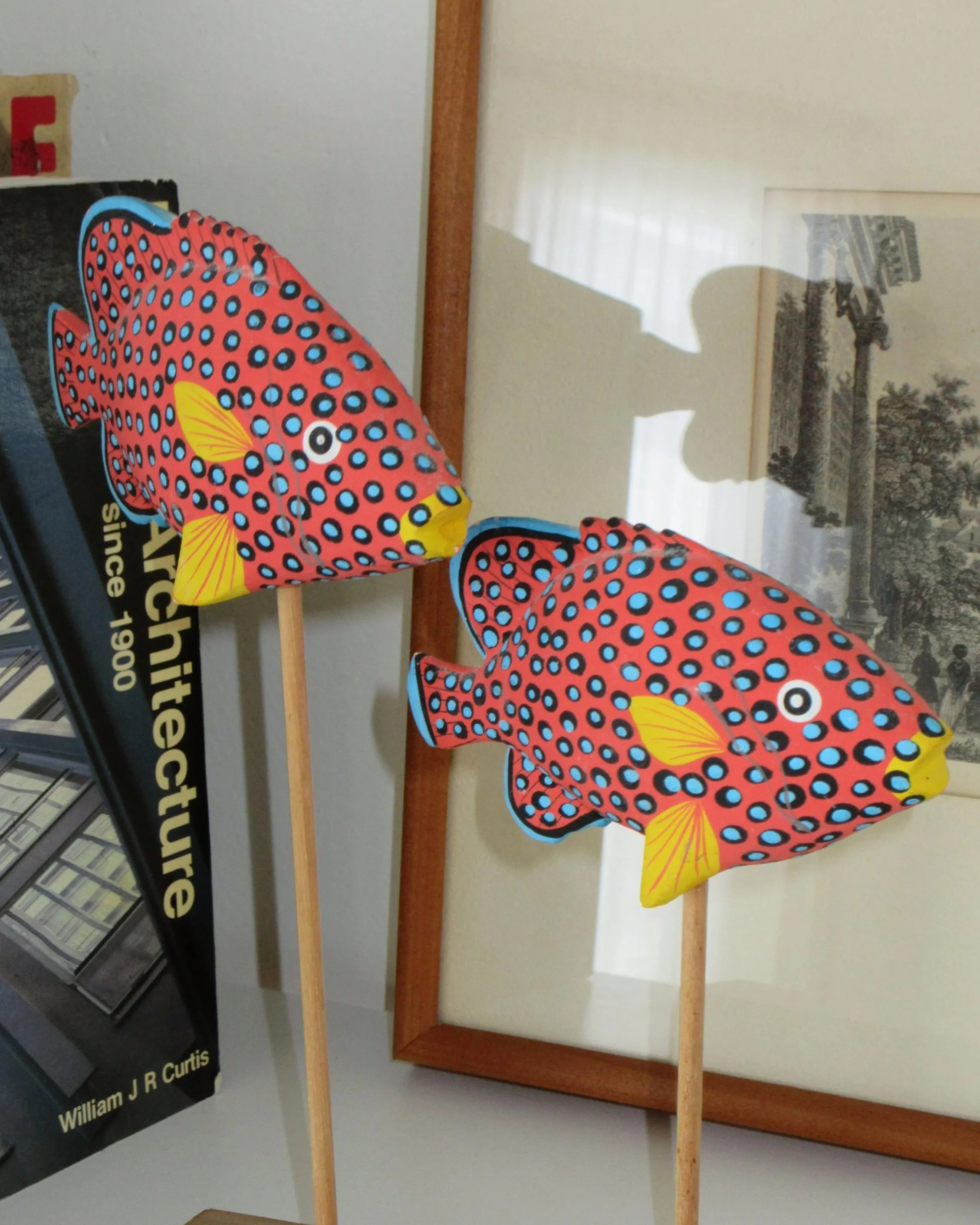 Two colorful fish-shaped paper lanterns with patterns of blue and black spots and yellow fins, mounted on wooden sticks, placed on a reflective surface with a framed picture and a book in the background.