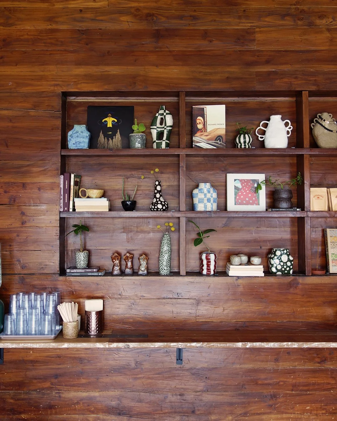 Wooden wall with a shelf holding various decorative vases, books, and small plants. On the counter below, there are cups, a container with wooden stirrers, and a metallic holder.