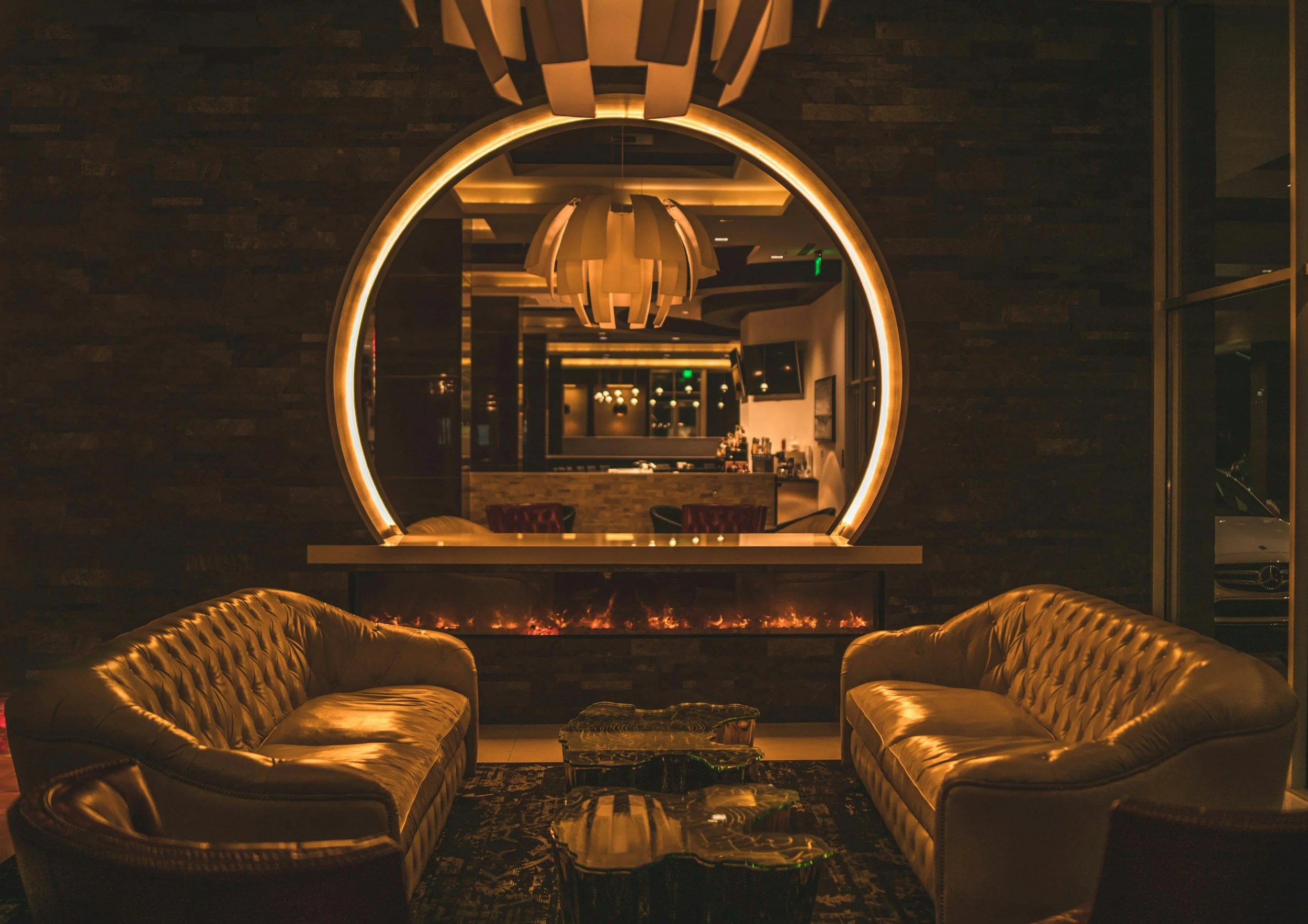 Stylish lounge area with two tan leather sofas facing each other, a black coffee table in between, behind which is a fireplace with a glowing fire, and a large circular mirror on the dark accent wall reflecting the room's warm lighting.