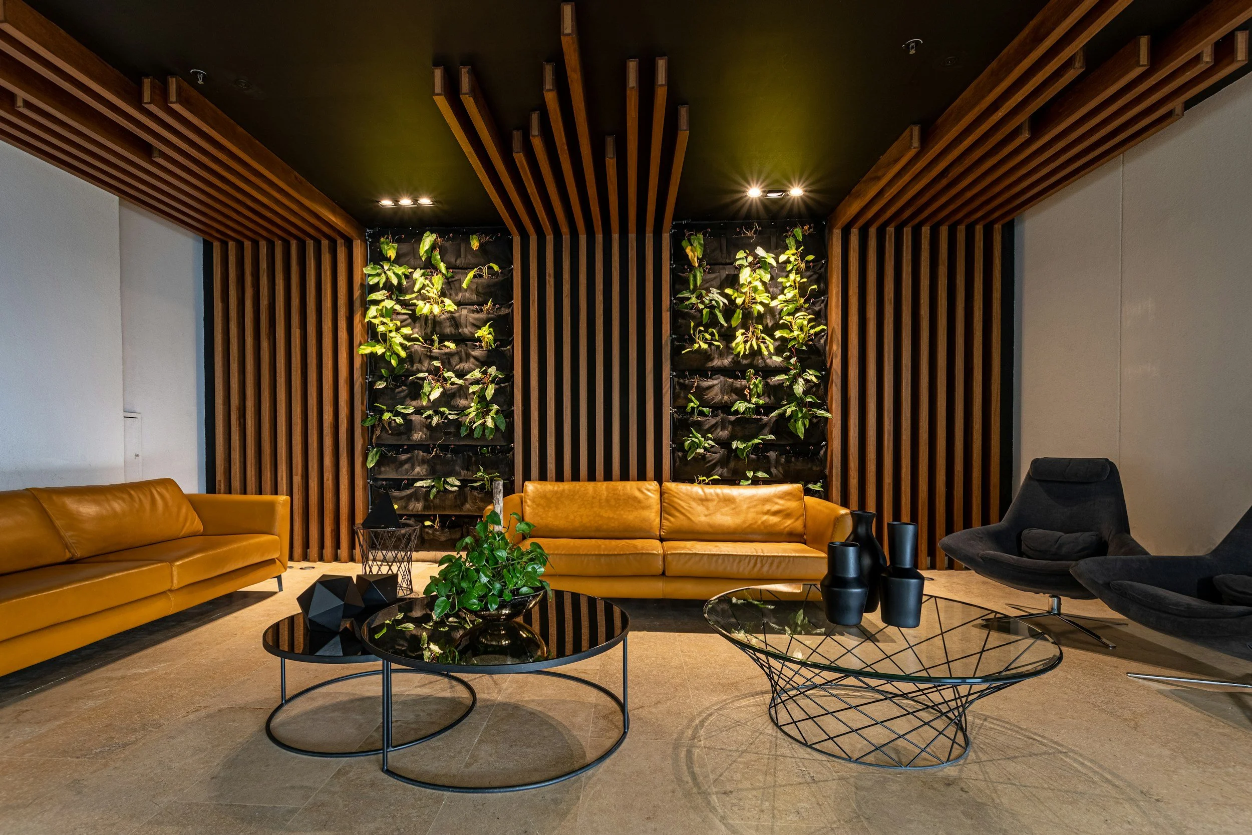 Modern living room with tan leather sofas, black wireframe coffee tables, and a vertical plant wall feature with green plants, black planters, and wooden accents on the ceiling and walls.