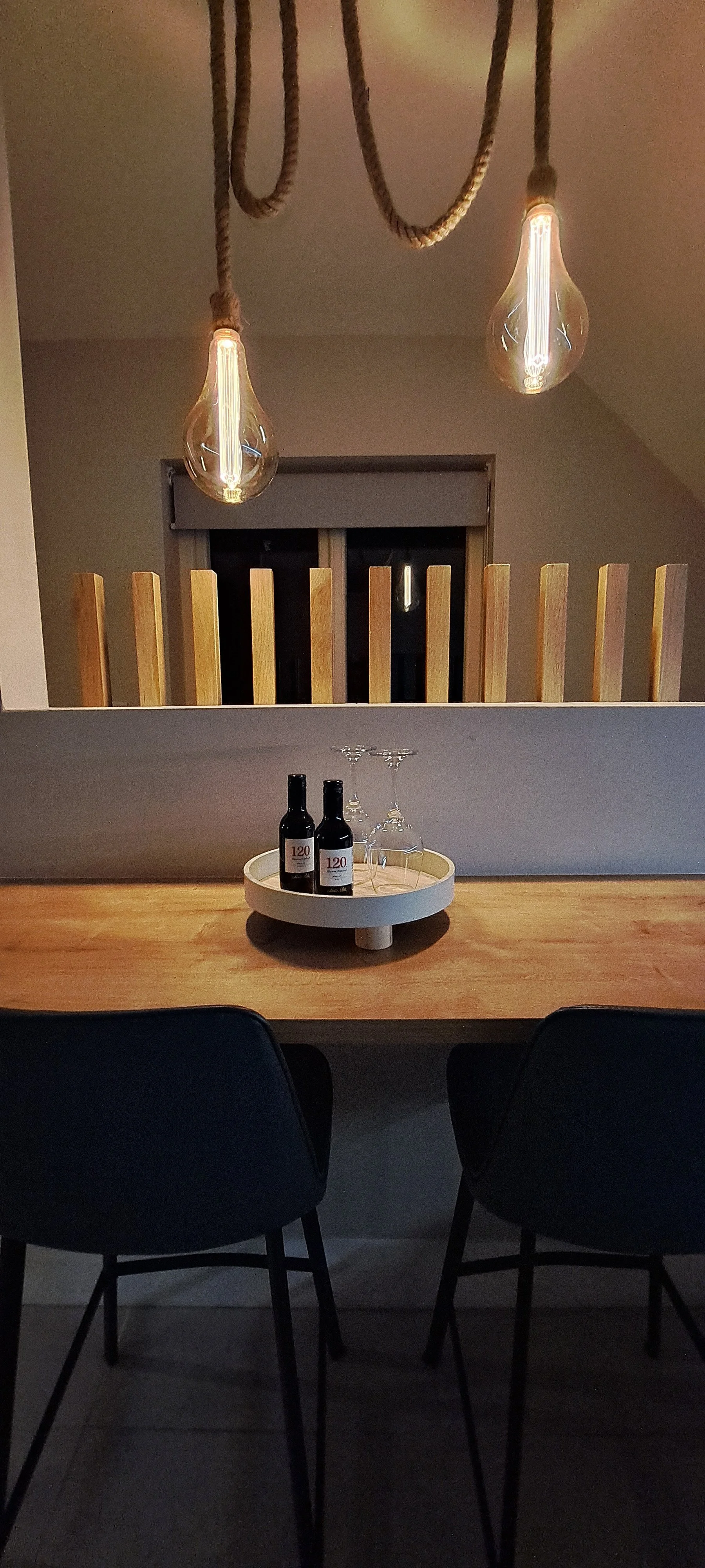 A modern dining area with two black chairs, a wooden table, two wine bottles, and two wine glasses on a round tray, illuminated by hanging light bulbs with ropes.