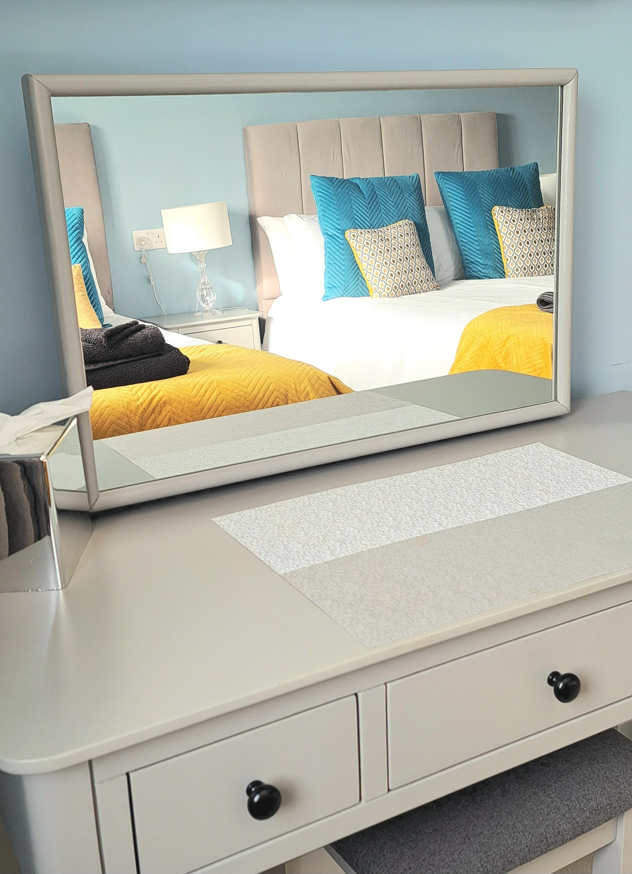 A mirror reflecting a bedroom with a bed that has a beige headboard, white bedding, blue pillows, and yellow pillows. A bedside table with a lamp and some folded blankets is visible. The furniture is white, and the wall behind the bed is painted ligh