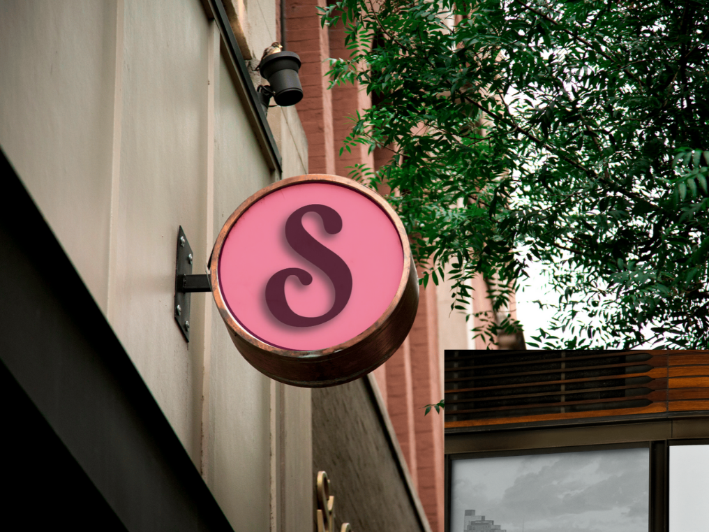 Signage attached to a building, looking up towards it with leaves in the background, the sign has a pink circle with maroon S with a shadow down to the left