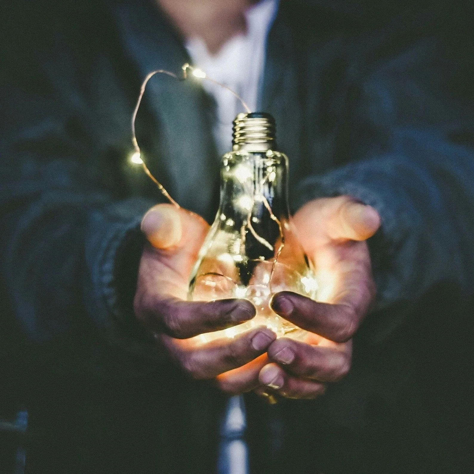 Person holding a lightbulb with a glowing string of fairy lights inside, creating an illuminated effect in a dark setting.