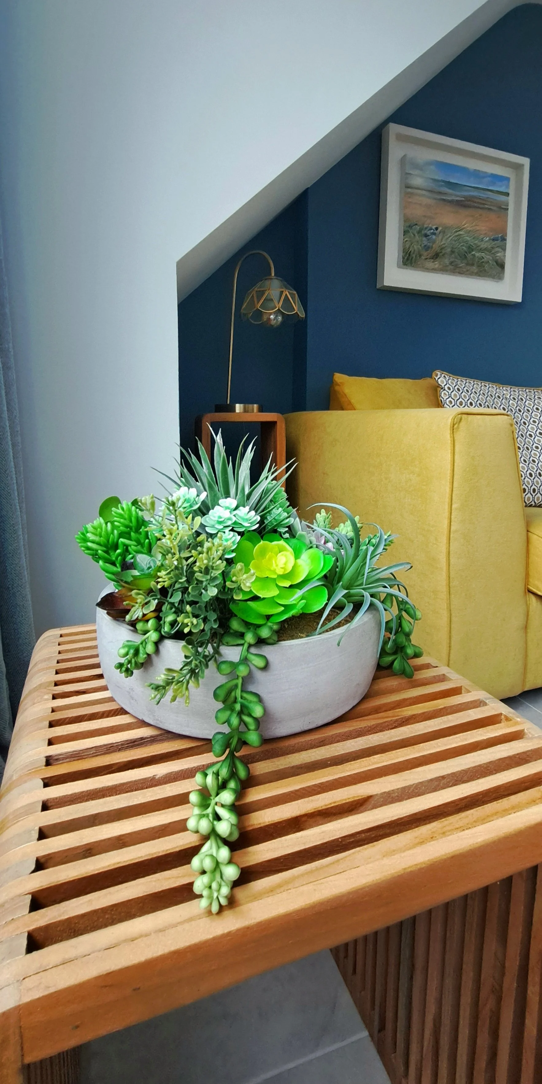 A decorative arrangement of artificial succulents in a gray bowl on a wooden slatted table, with a yellow couch, a framed painting, and a floor lamp in the background.