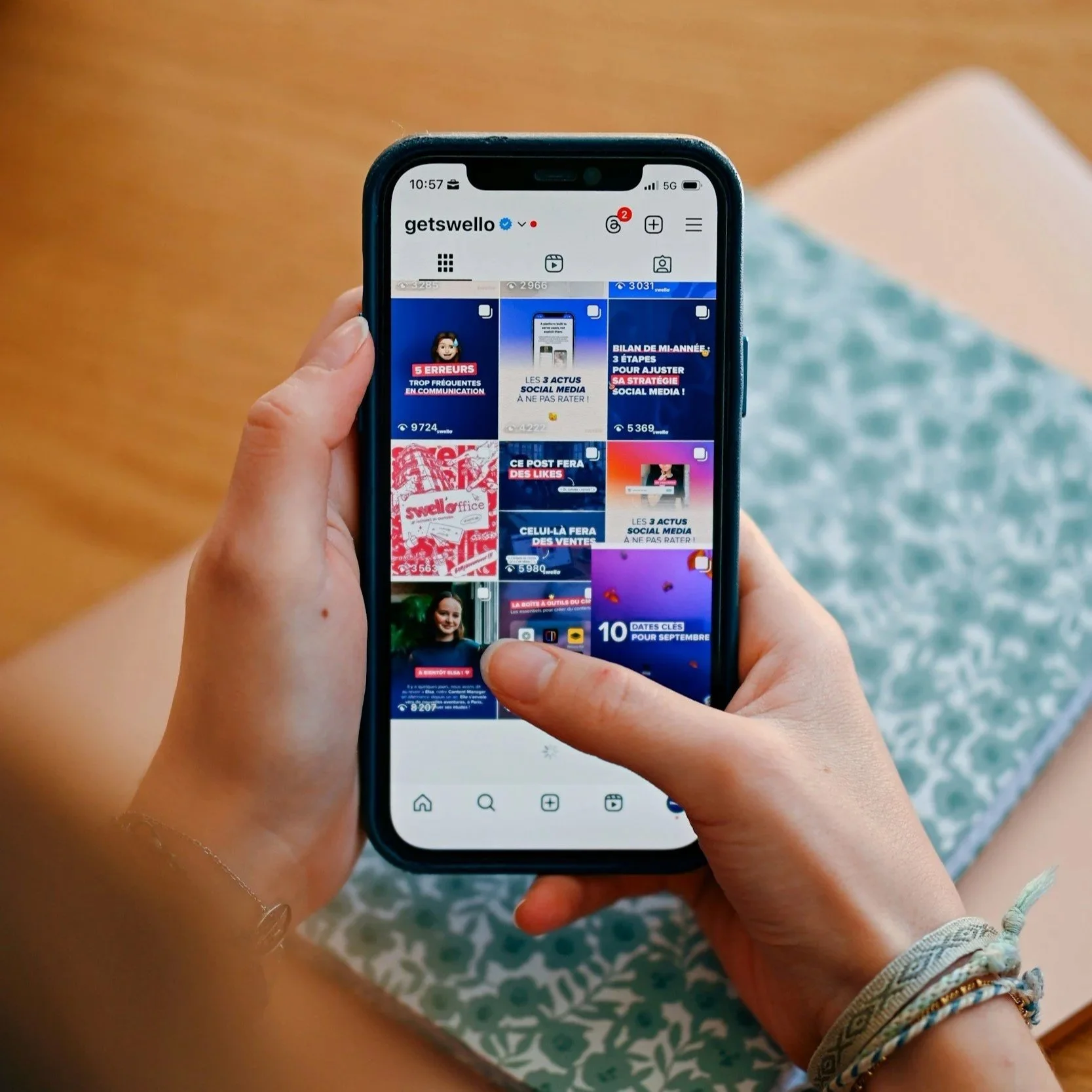 Close-up of a person holding a smartphone displaying an Instagram profile with a grid of social media posts, featuring primarily blue and red graphics, and a background of a patterned blanket and wooden floor.