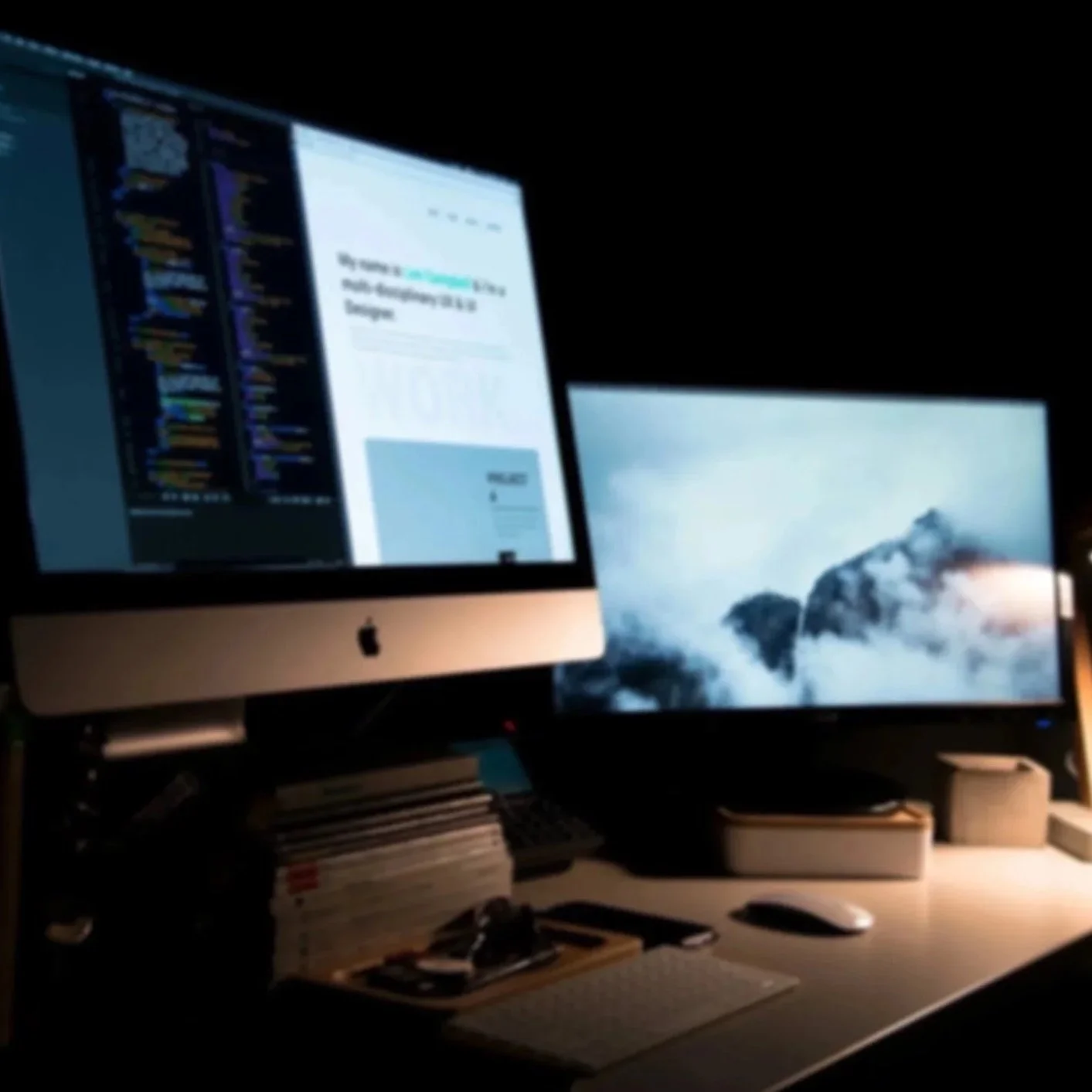 Desk with iMac computer displaying code and design software, dual monitors, and various office supplies in a dark room