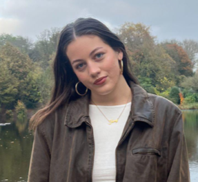 Young woman with dark hair, wearing hoop earrings and a brown jacket, standing outdoors near a body of water with trees in the background.