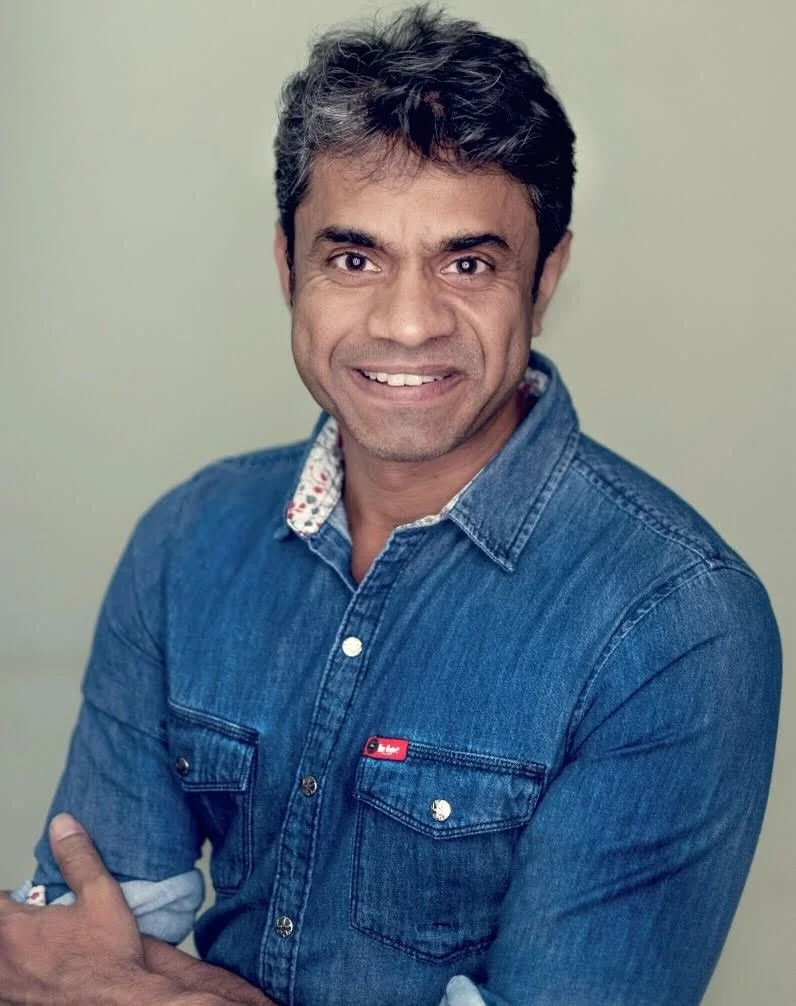 A smiling man with dark hair and tan skin, wearing a denim shirt with a floral collar, standing against a plain wall.