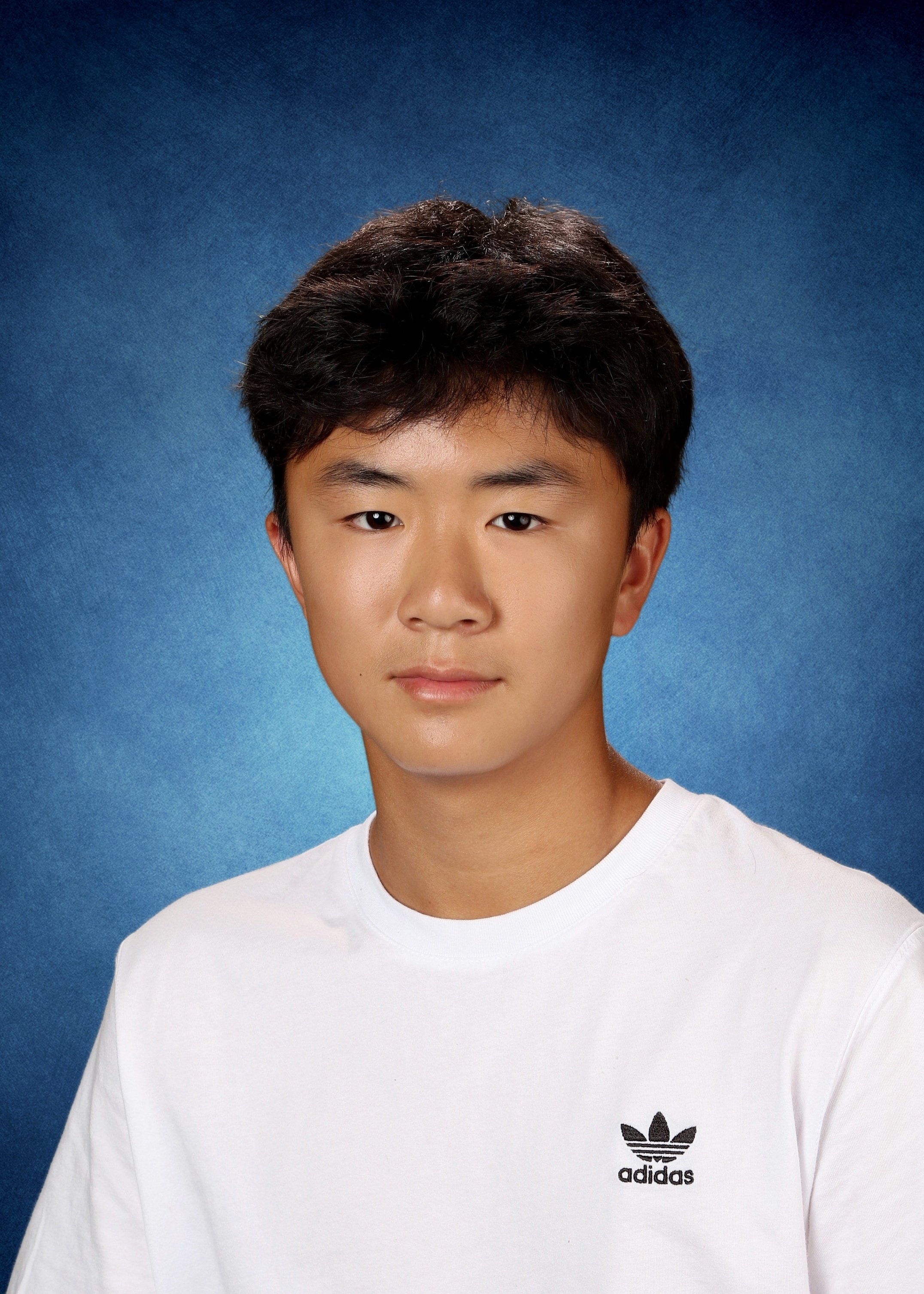 Portrait of a young man with short dark hair, wearing a white Adidas T-shirt, against a blue background.