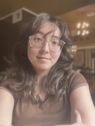A woman with glasses and wavy hair, wearing a brown shirt, taking a selfie in an indoor setting.