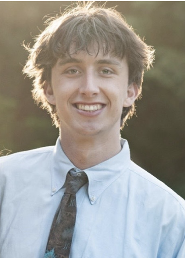 Young man with curly hair, smiling, wearing a light blue dress shirt and a dark tie outdoors.