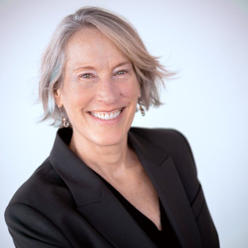 A woman with short gray hair smiling, wearing a black blazer and earrings, against a plain white background.