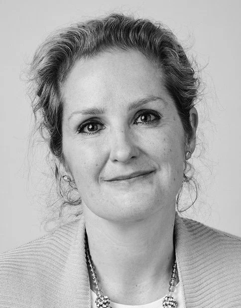 Black and white portrait of a woman with wavy hair, wearing a light blazer and a necklace, smiling softly at the camera.