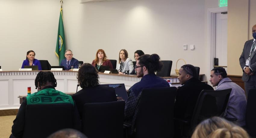 A group of people sitting around a conference table in a formal meeting room, with some taking notes and others listening, and a panel of officials or speakers at the front of the room.