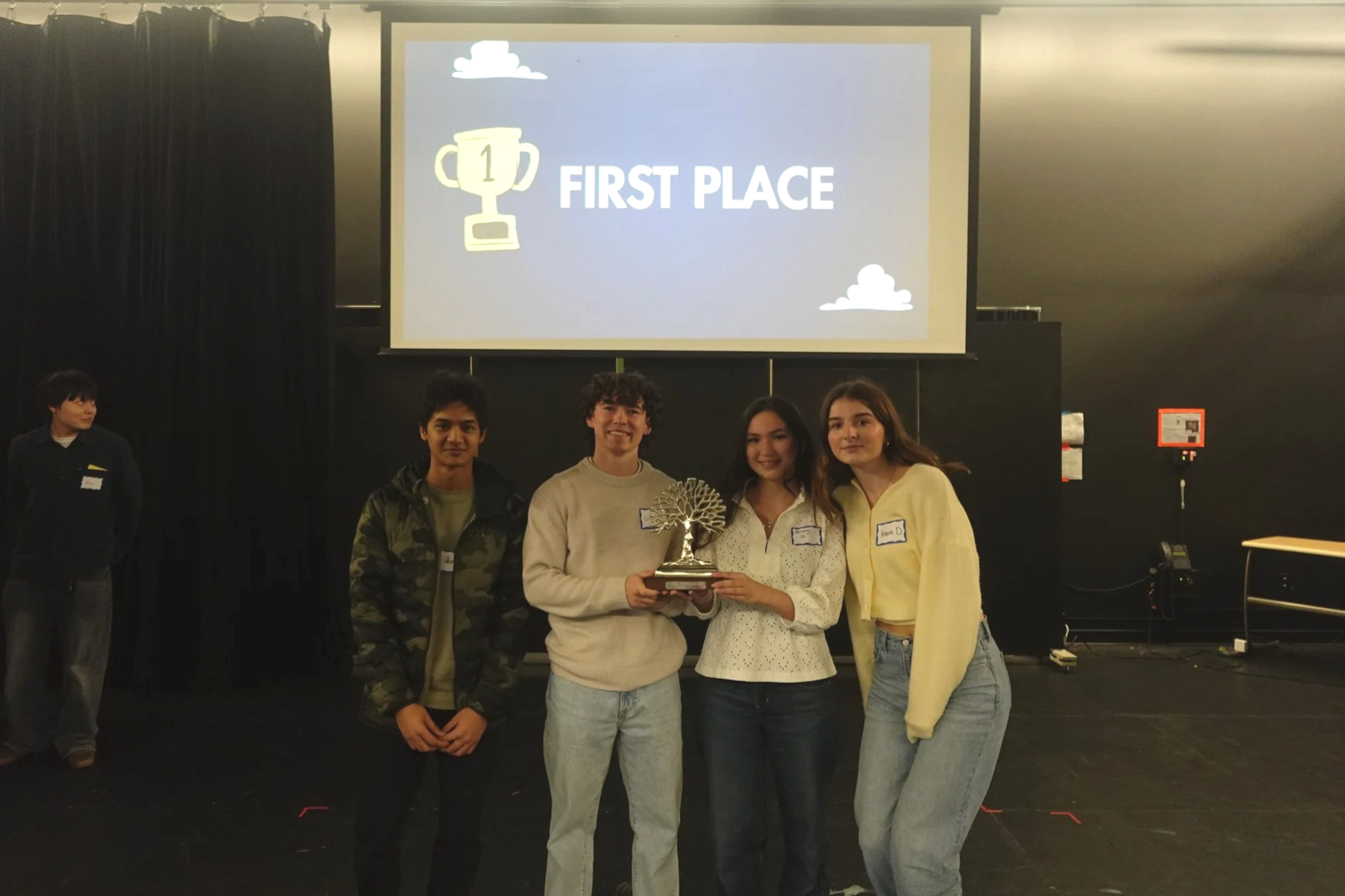 Group of four young people standing together on stage, holding a trophy with a tree design, in front of a large screen displaying 'FIRST PLACE' and a trophy icon. A person stands to the left, and the background is dark with some equipment visible.