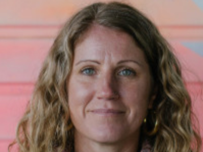 A woman with curly hair and light skin, smiling lightly, with a pink background.