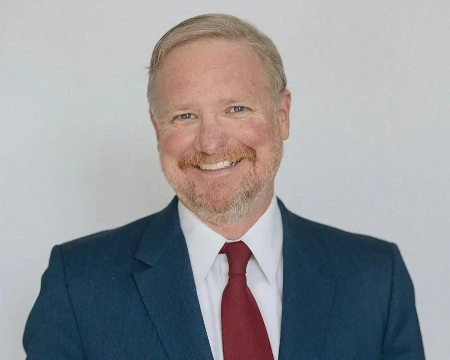 Portrait of a smiling middle-aged man with light hair and a beard, wearing a navy suit, white shirt, and red tie, against a plain light background.
