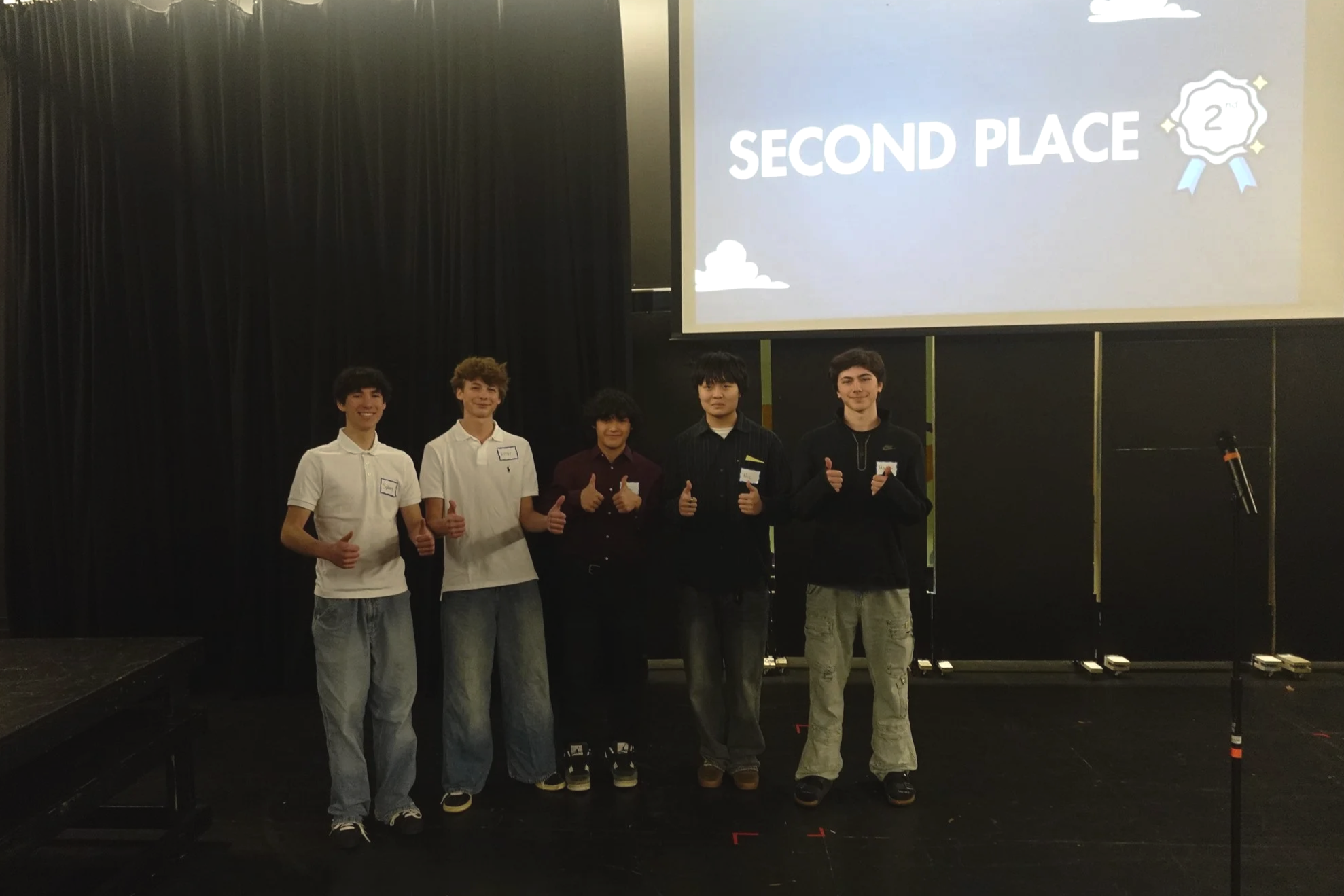 Five young men standing on stage in front of a large screen displaying 'Second Place' with shield and ribbon icons