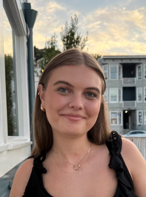 A young woman with long brown hair, blue eyes, and light skin, smiling outdoors during sunset. She is wearing a black sleeveless top, gold earrings, and layered necklaces. Background includes a modern building, trees, and a partly cloudy sky.