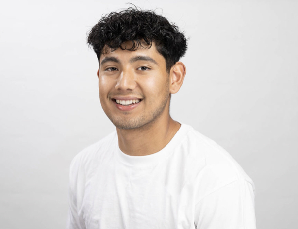 A smiling young man with dark, curly hair wearing a white t-shirt against a plain light gray background.