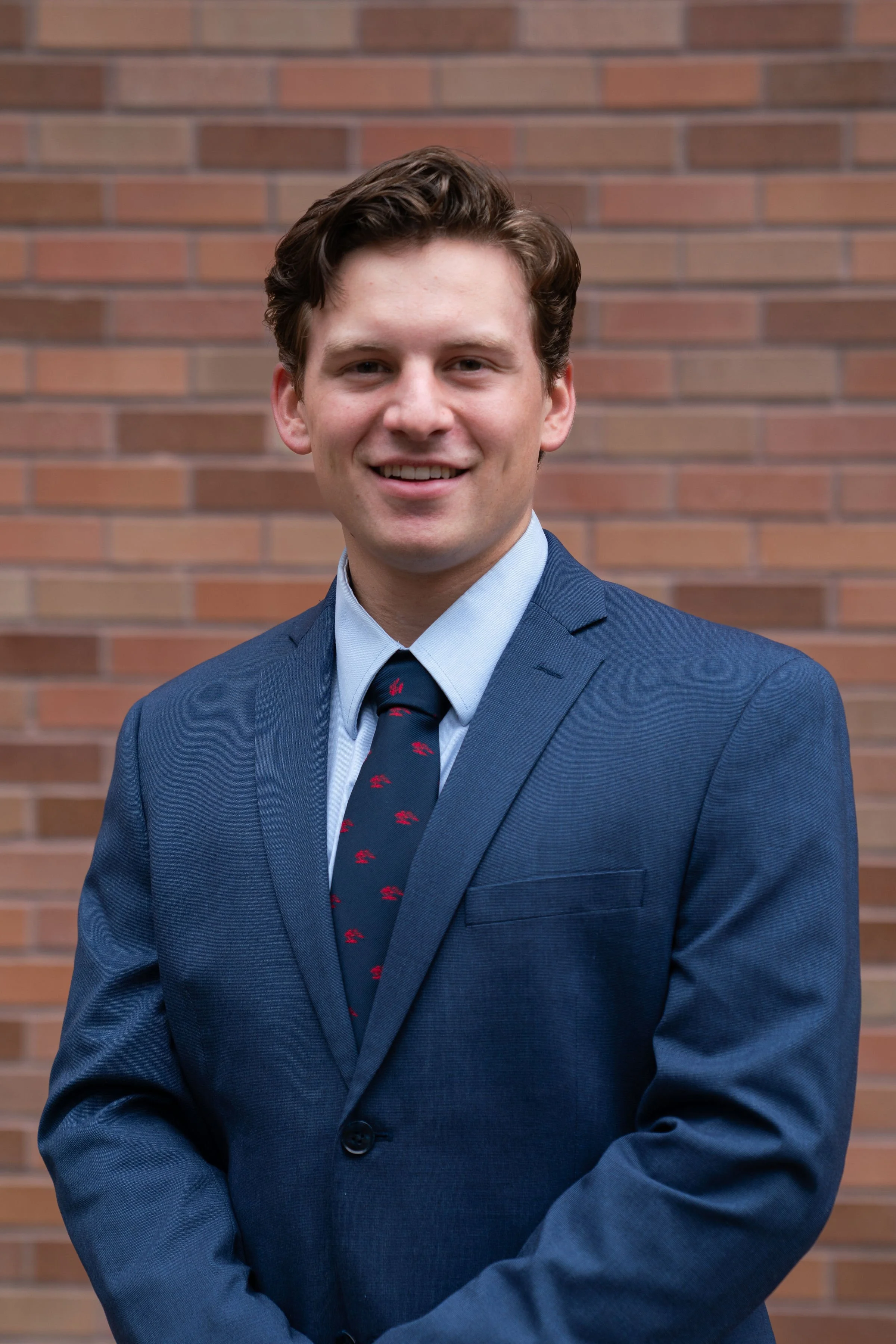 A young man in a navy blue suit, light blue shirt, and patterned tie standing in front of a brick wall, smiling at the camera.
