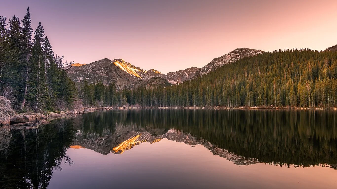 Serene mountain lake surrounded by pine trees with snowy peaks in the background during sunset.