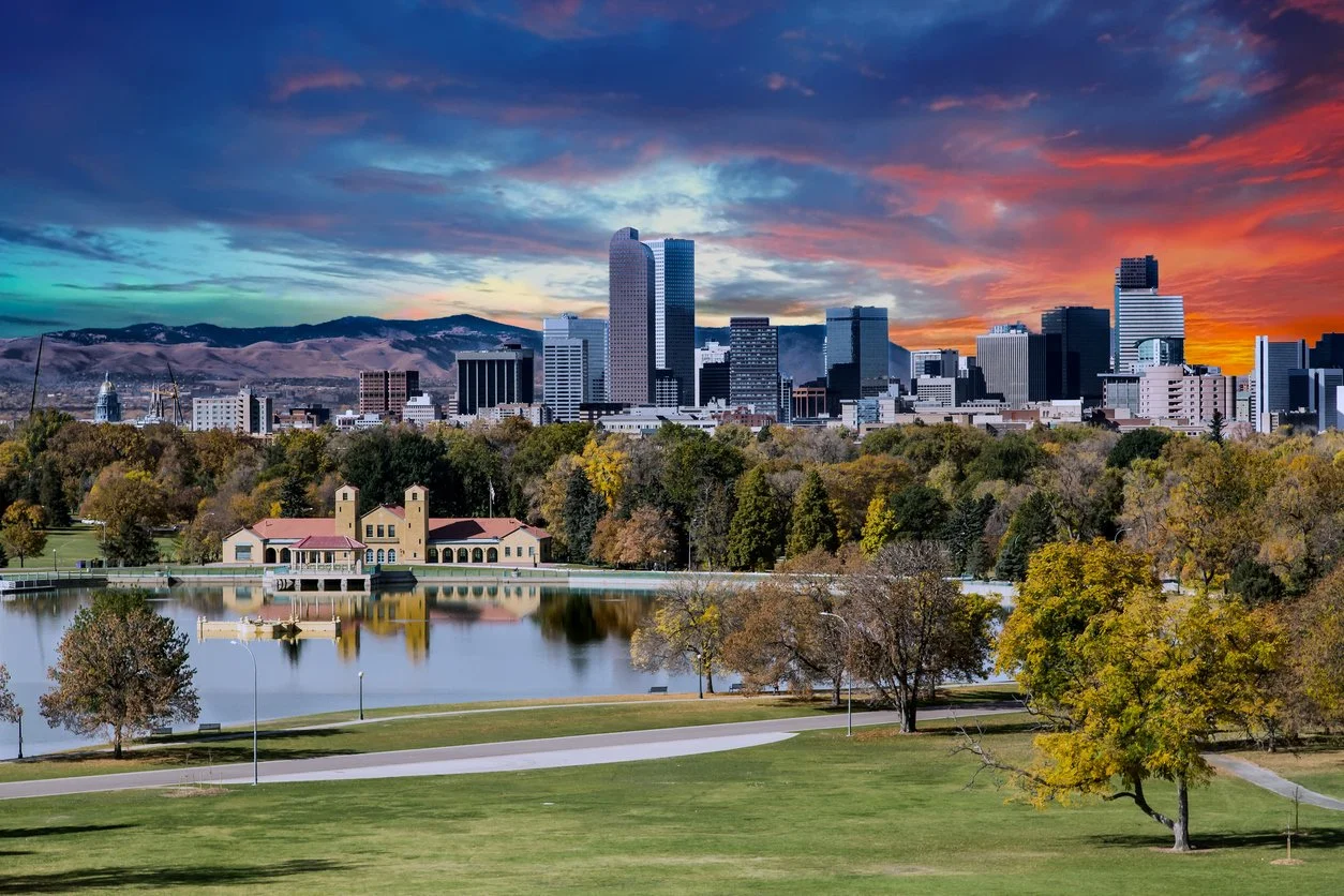The image shows the city skyline of Oklahoma City with tall skyscrapers in the background, a park with green grass and trees in the foreground, and a small lake with a historic building beside it. The sky is colorful with shades of blue, orange, pink, and purple during sunset.