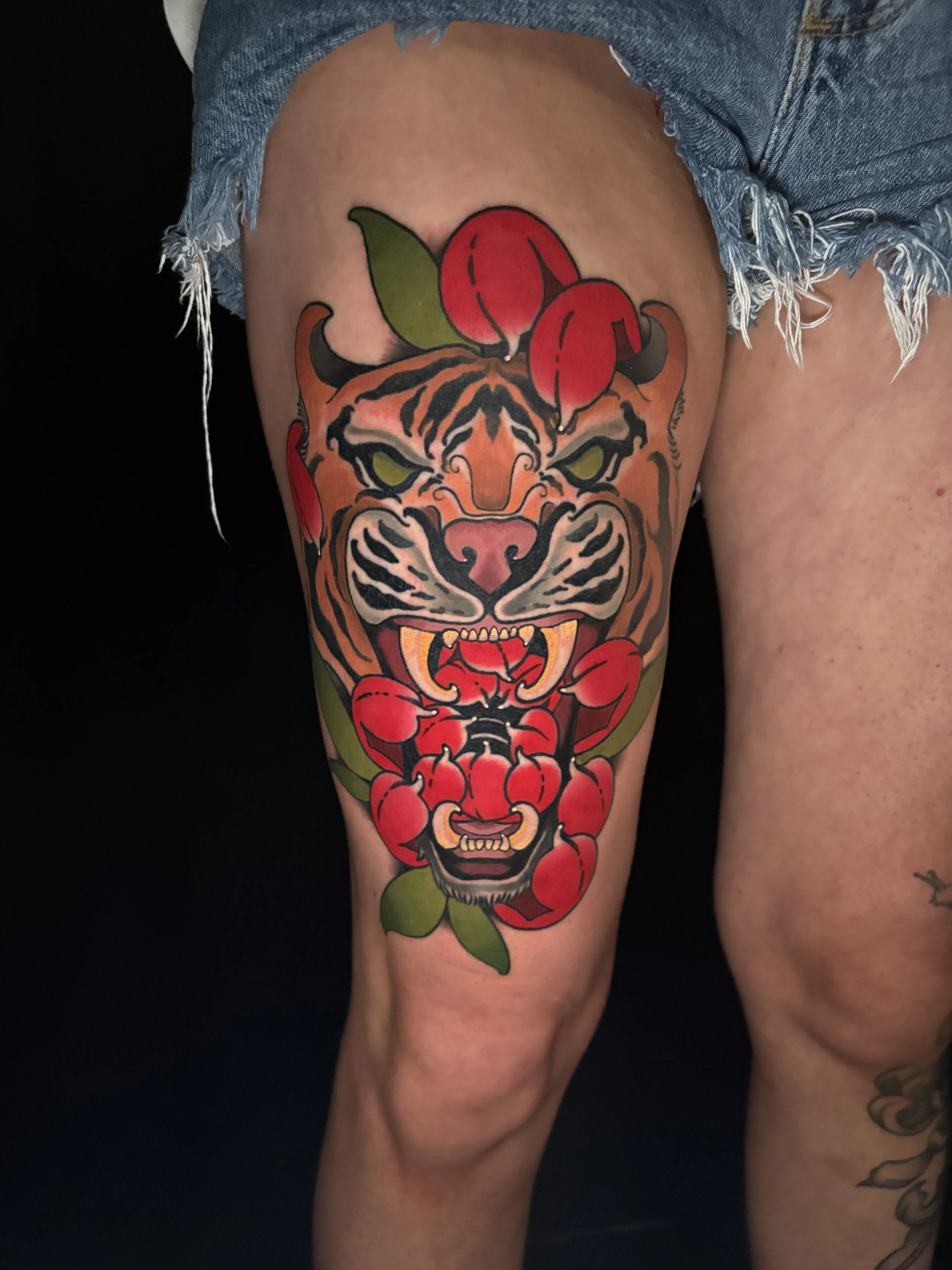 Tattoo of a fierce tiger face with green eyes, surrounded by red chrysanthemum and green leaves on a person's thigh.