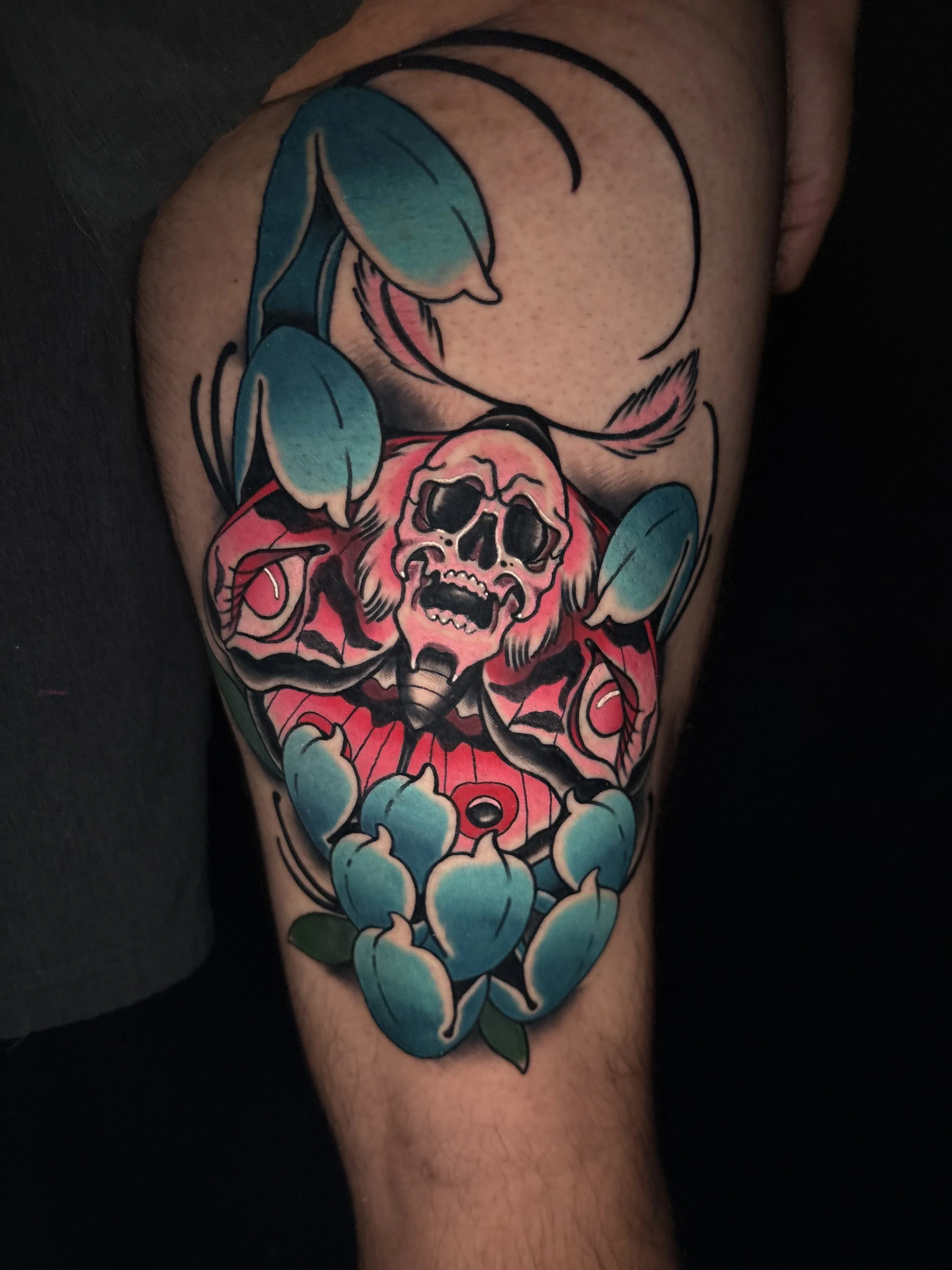 Tattoo of a colorful skull moth with pink and black details, surrounded by blue-green chrysanthemum, on a person's thigh.