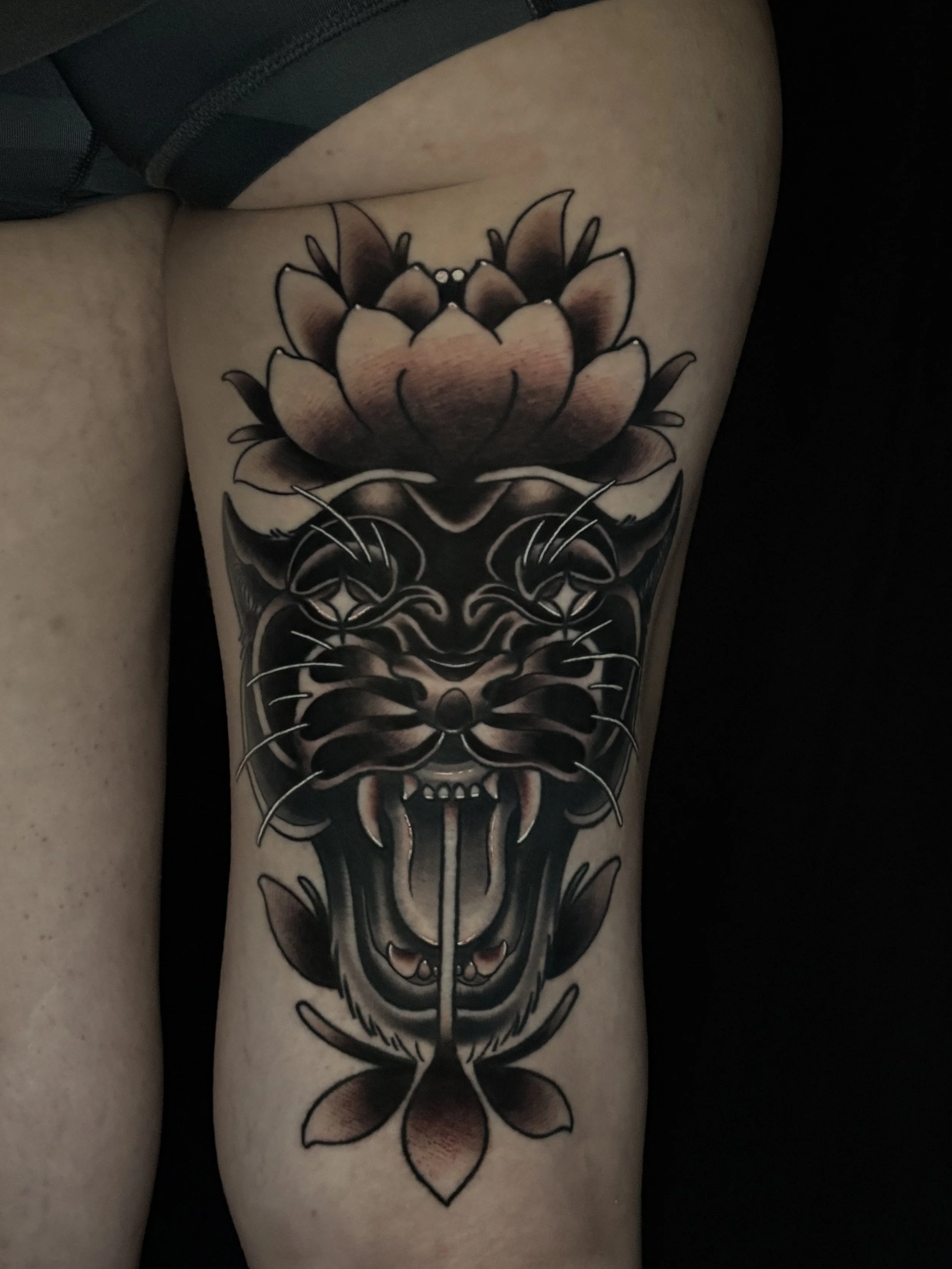 A tattoo of a detailed, stylized panther face with black, gray, and flesh-colored shading on a person's thigh.