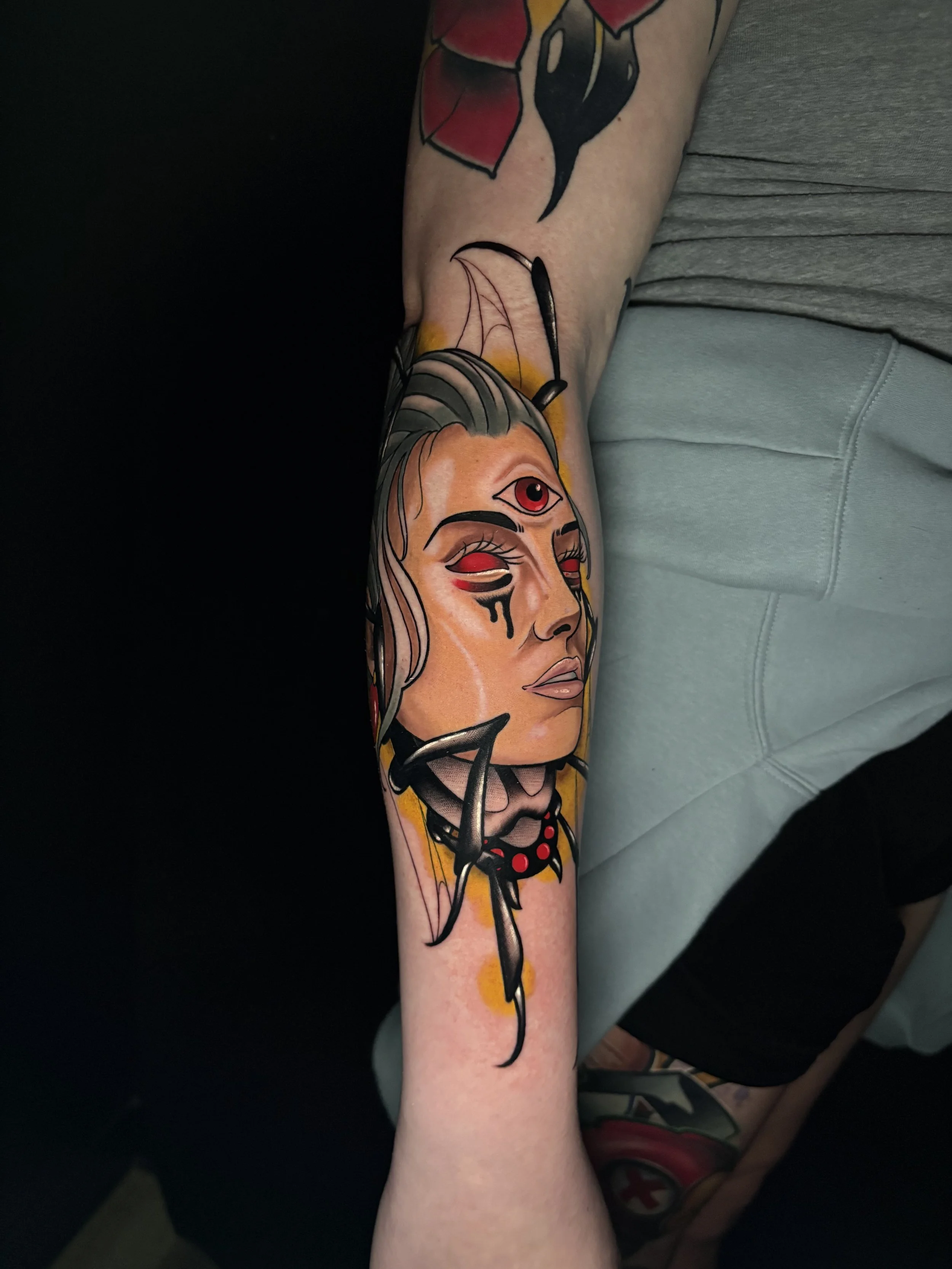 Colorful tattoo of a woman with a third eye on her forehead, with black and red eye makeup, long hair, and decorative elements on her face, on a person's forearm.