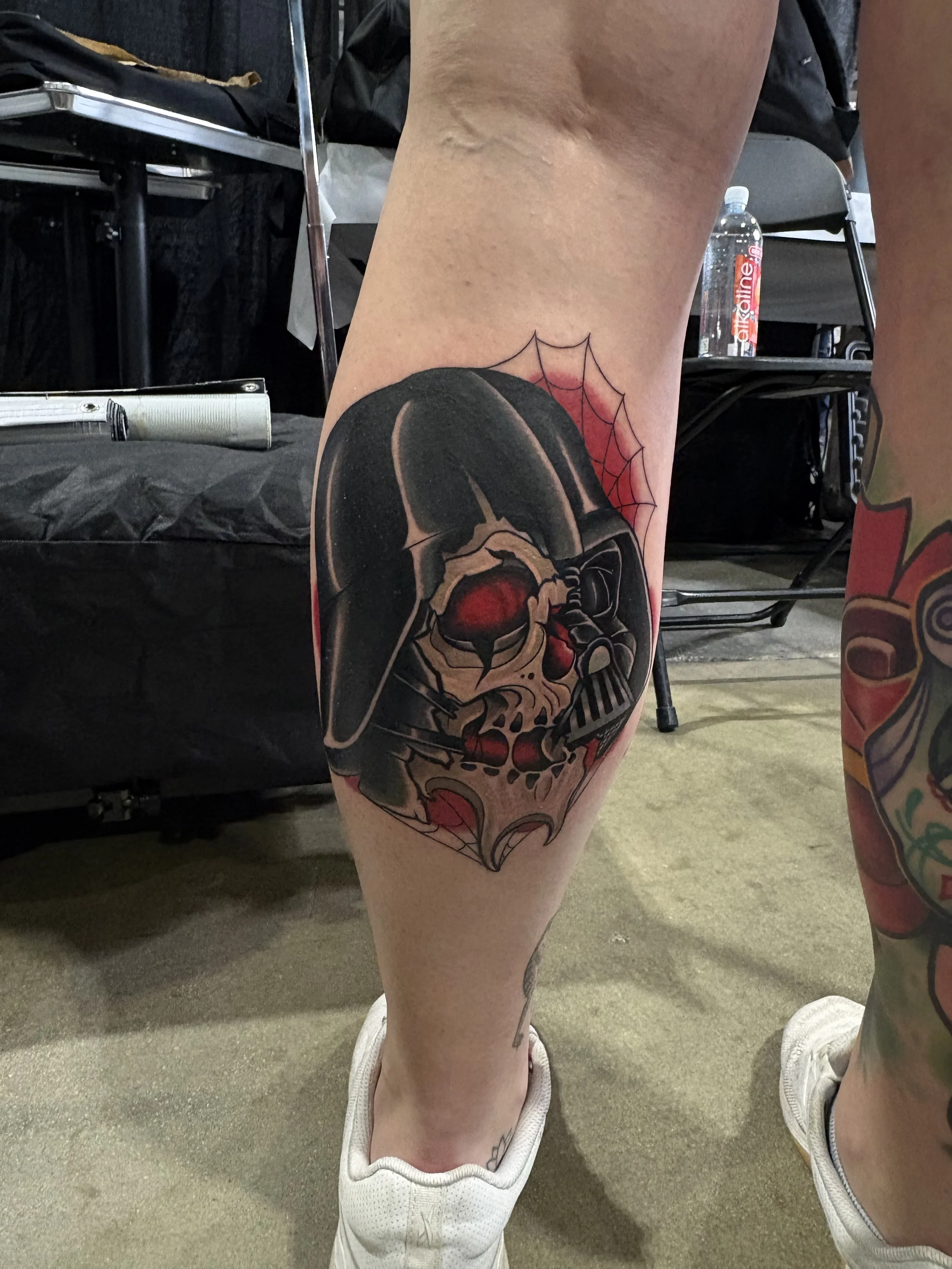 A person's leg with a tattoo of a star wars darth vader skull with red eyes and a spider web on a black background.